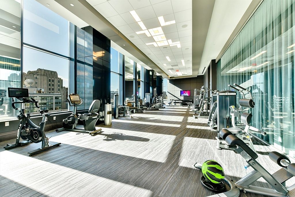This is a well-equipped home gym featuring large windows with city views, various cardio machines like stationary bikes and treadmills, and weight training equipment. The space is brightly lit with modern ceiling fixtures and has a clean, contemporary design with a neutral color palette, making it an appealing amenity for potential residents.