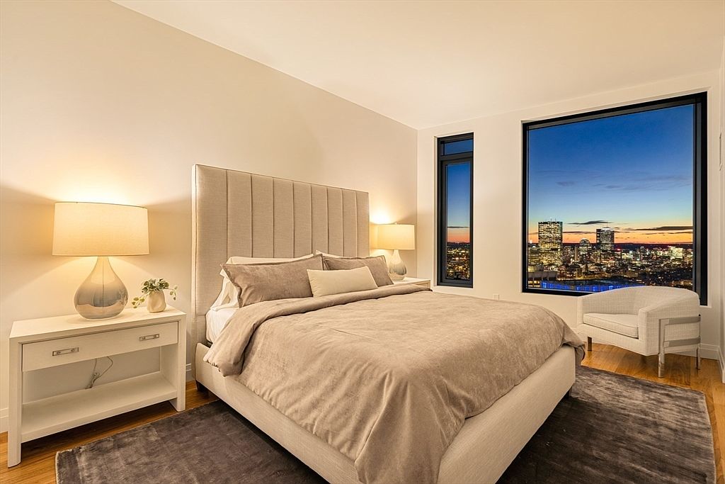 This is a primary bedroom featuring a large bed with a padded headboard, neutral-toned bedding, and two nightstands with lamps. Large windows offer a view of the city skyline at dusk, and a modern armchair sits near the window. The room has a sophisticated and calming atmosphere.