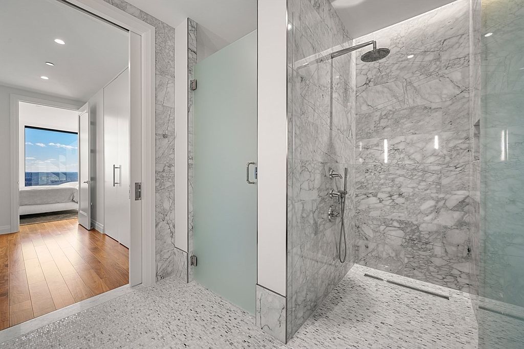This is a modern primary bathroom featuring a walk-in shower with marble walls and a glass enclosure. The shower has a rainfall showerhead and sleek fixtures. The bathroom floor is tiled with small mosaic tiles, and there is an open doorway leading to the bedroom, which has hardwood floors and a large window with a view.