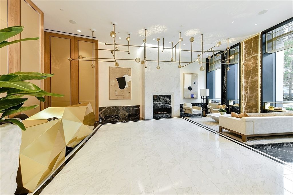This is an interior shot of a luxurious lobby or hallway, featuring a polished white marble floor with black accents. A modern, geometric gold reception desk sits to the left, complemented by abstract art and a unique chandelier. The space includes a seating area with a neutral-toned sofa and chairs, creating an inviting and sophisticated atmosphere.