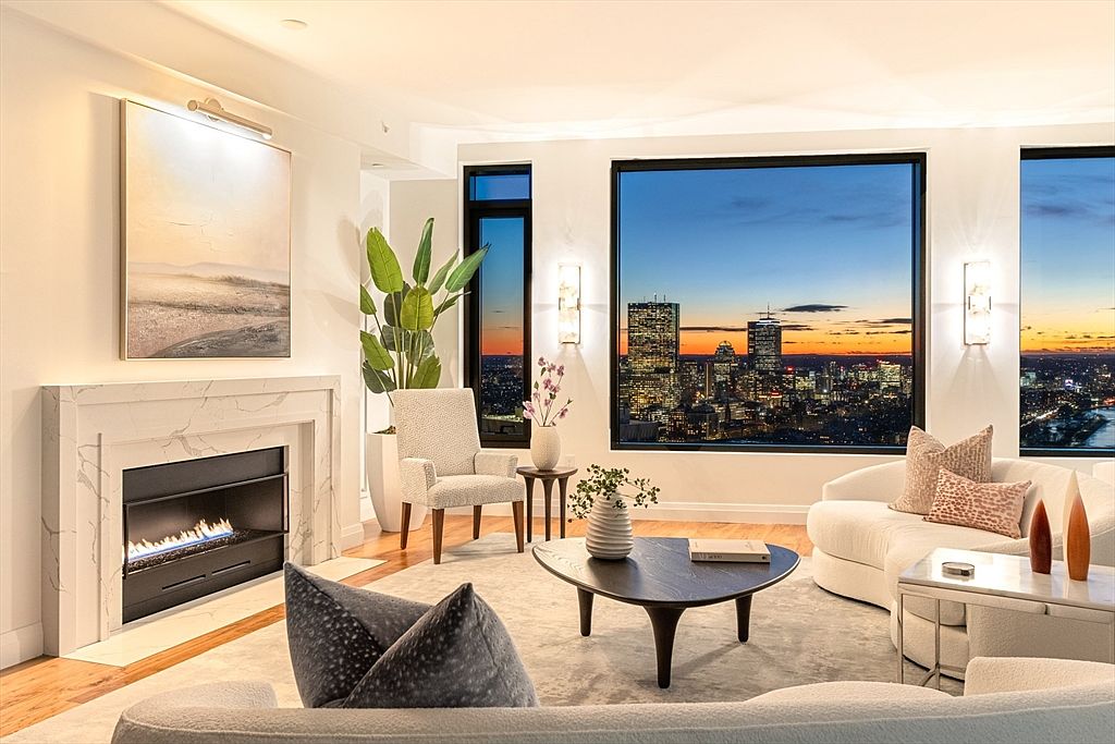 This is a luxurious living room featuring a modern fireplace with a marble surround and a large abstract painting above it. The room is furnished with elegant, curved sofas and a unique coffee table, complemented by a stunning cityscape view through large windows. The overall impression is one of sophistication and comfort, perfect for upscale living.
