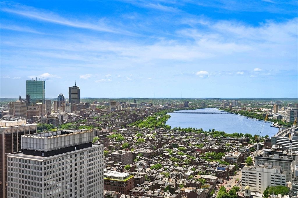 This aerial view showcases a sprawling cityscape with a prominent river running through it. The city features a mix of high-rise buildings and dense residential areas, all set against a backdrop of a clear blue sky with scattered clouds. The image provides a comprehensive overview of the urban landscape, highlighting its scale and layout.