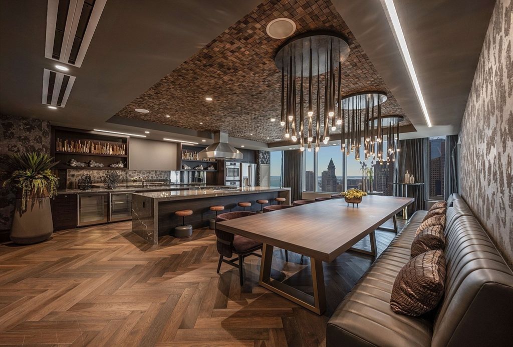 This is a luxurious kitchen and dining area featuring a large island with bar seating, custom cabinetry, and high-end appliances. The dining area includes a long wooden table with a built-in banquette and modern chandelier lighting. The herringbone wood flooring and textured ceiling add to the sophisticated ambiance, while large windows offer city views.