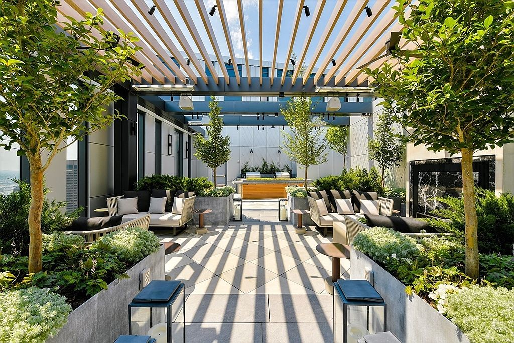This image showcases a luxurious outdoor patio or balcony area, featuring comfortable lounge seating arranged around lush greenery and trees in planters. A modern pergola provides shade, casting geometric shadows on the tiled floor. The space is designed for relaxation and entertainment, offering a sophisticated urban oasis.