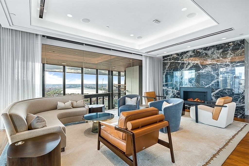 This is a luxurious living room featuring a curved sofa, stylish armchairs, and a modern fireplace with a marble surround. Large windows offer panoramic city views, and the room is decorated in neutral tones with pops of color. The overall impression is one of sophistication and comfort.