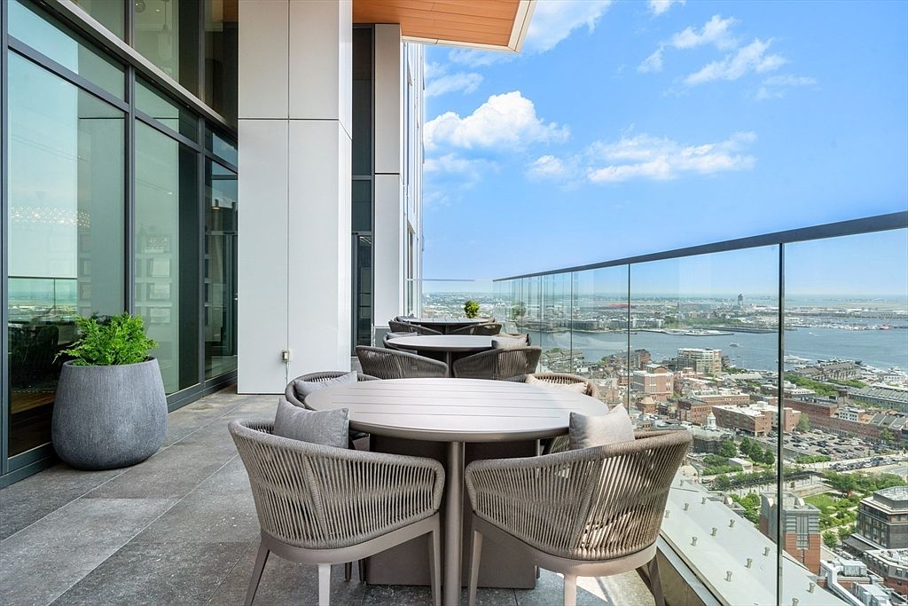 This image showcases a luxurious outdoor balcony area, featuring modern patio furniture including round tables and woven chairs with cushions. The balcony offers stunning panoramic views of the city and waterfront, framed by a sleek glass railing. A large potted plant adds a touch of greenery to the sophisticated outdoor space.