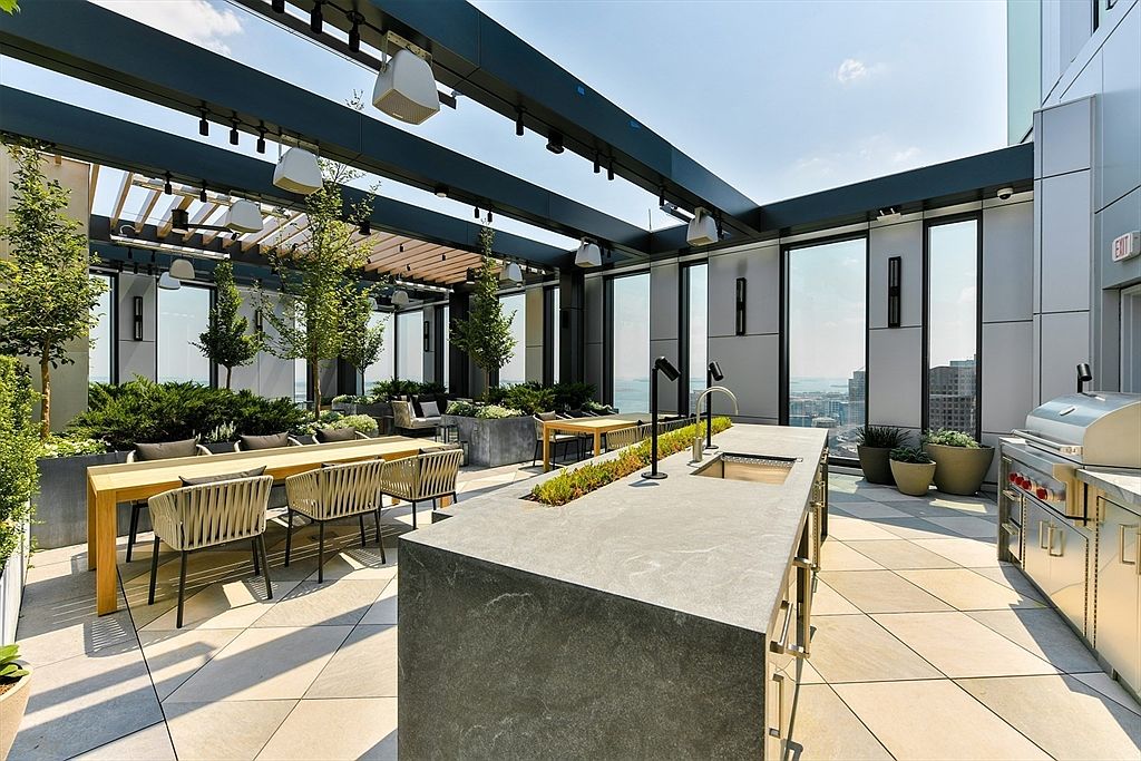 This image showcases a luxurious rooftop patio or balcony, designed for outdoor living and entertaining. It features a built-in outdoor kitchen with a grill and countertop space, a long dining table with stylish chairs, and comfortable lounge seating surrounded by greenery. The space is covered by a modern pergola structure, providing shade and architectural interest, while offering city views.