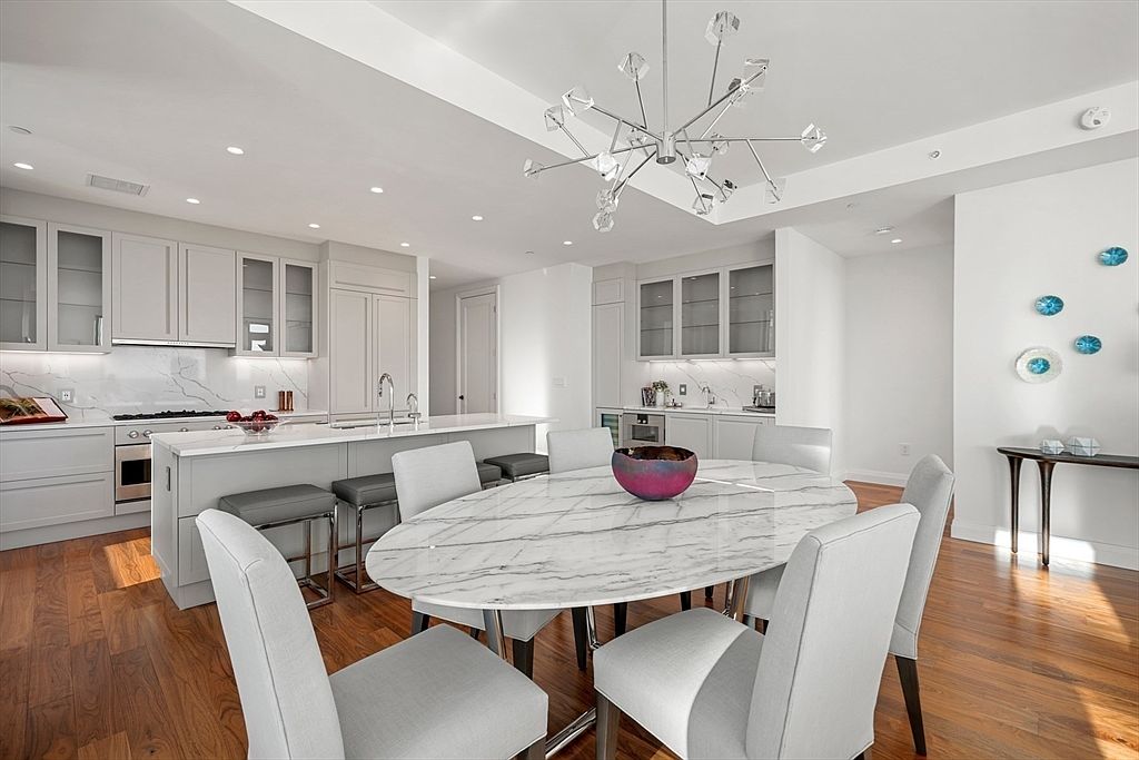 This is a bright and modern kitchen and dining area featuring white cabinetry, marble countertops, and stainless steel appliances. The dining area includes an oval marble table with light gray chairs and a unique chandelier overhead. The hardwood floors add warmth to the space, creating an inviting and luxurious atmosphere.