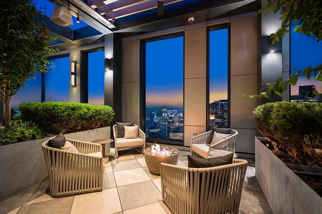 This image showcases a luxurious outdoor balcony or patio area, featuring comfortable seating around a modern fire pit. The space is adorned with lush greenery in planters, and large windows offer stunning city views at dusk. The overall impression is one of sophisticated urban living and relaxation.