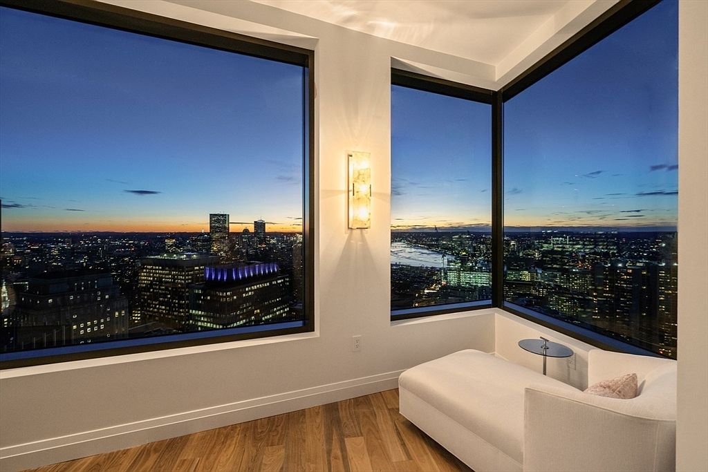 This is an interior shot of a modern living room featuring floor-to-ceiling windows that offer a panoramic city view at dusk. The room is minimally furnished with a white chaise lounge and a small side table, emphasizing the view and the natural light. An elegant wall sconce provides additional lighting, enhancing the room's sophisticated ambiance.