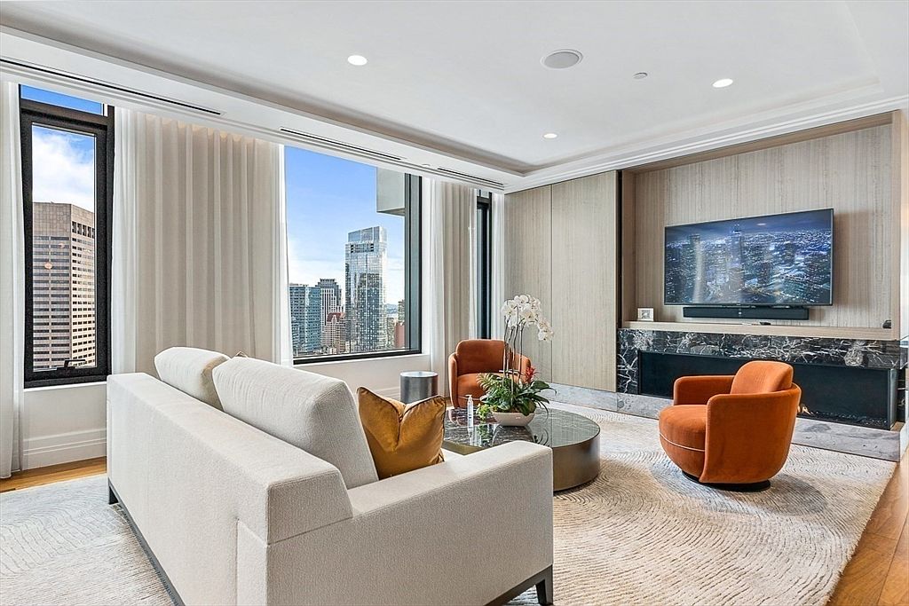 This is a luxurious living room featuring a modern design with high-end finishes. The room includes a comfortable white sofa, two orange armchairs, and a round coffee table, all set on a patterned rug. Large windows offer stunning city views, and a sleek fireplace with a mounted TV adds a focal point to the space.