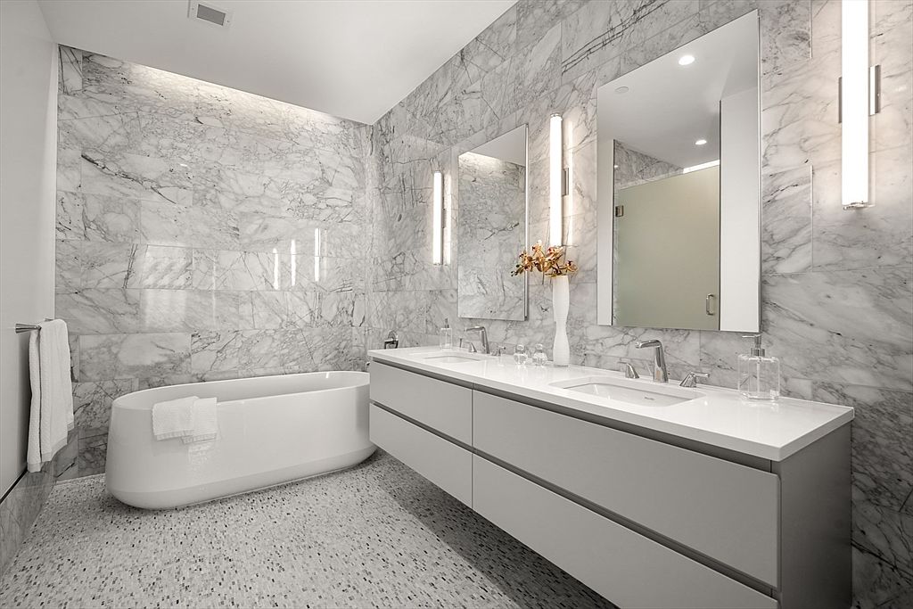 This is a luxurious primary bathroom featuring marble walls and flooring. A modern, freestanding bathtub sits adjacent to a double vanity with a white countertop and sleek gray cabinetry. The room is illuminated by vertical light fixtures flanking the mirrors, creating a spa-like ambiance.