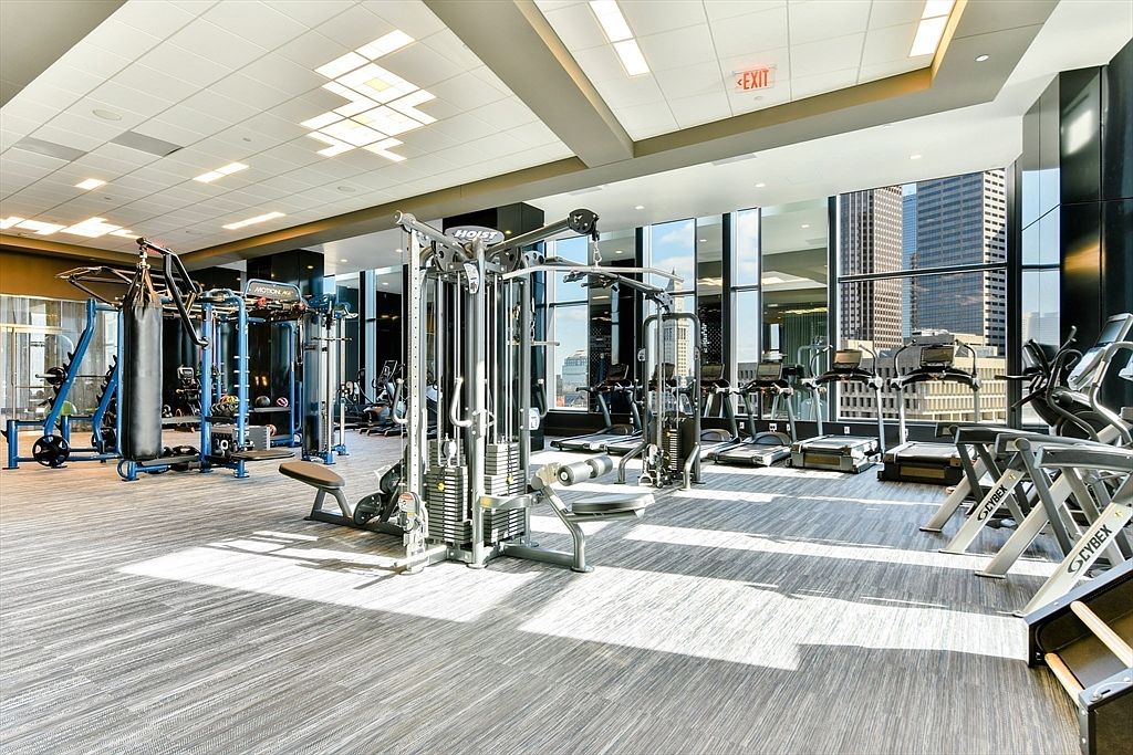 This is a well-equipped home gym featuring various exercise machines, including treadmills, weight machines, and a punching bag. Large windows offer a view of the city skyline, providing natural light and an inspiring workout environment. The flooring is a neutral-toned carpet, and the overall impression is modern and functional.