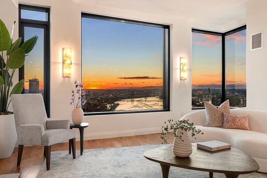 This is a beautifully staged living room with a modern aesthetic. Large windows offer stunning city views at sunset, complemented by elegant wall sconces. The room features a comfortable armchair, a curved sofa with decorative pillows, and a stylish coffee table, all set on a light-colored rug over hardwood floors.