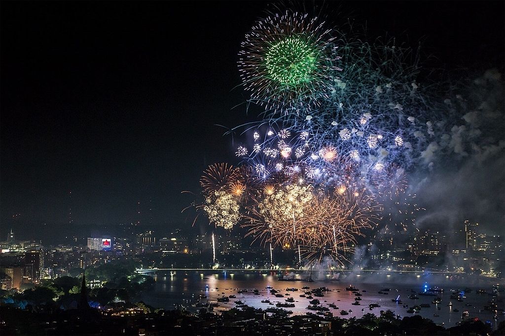 This aerial view captures a vibrant fireworks display over a city waterfront. The cityscape is illuminated by the colorful bursts, reflecting off the water where numerous boats are gathered. The scene conveys a sense of celebration and community, highlighting the city's lively atmosphere and scenic location.