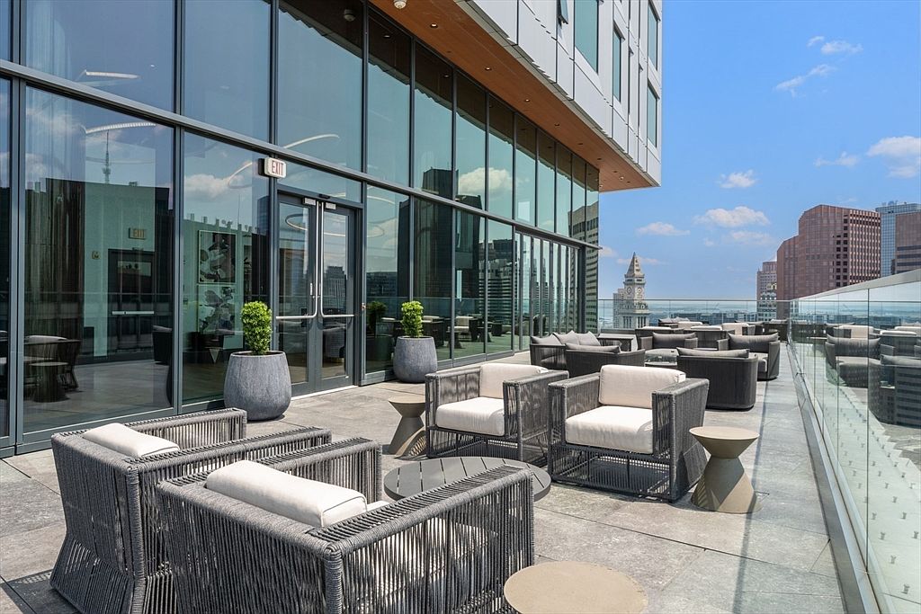 This image showcases a luxurious outdoor patio or balcony area, likely part of a high-end apartment or condominium. The space is furnished with modern, woven-frame seating arrangements and small tables, creating an inviting lounge area. The patio features a glass railing offering unobstructed city views, enhancing the appeal of the property.