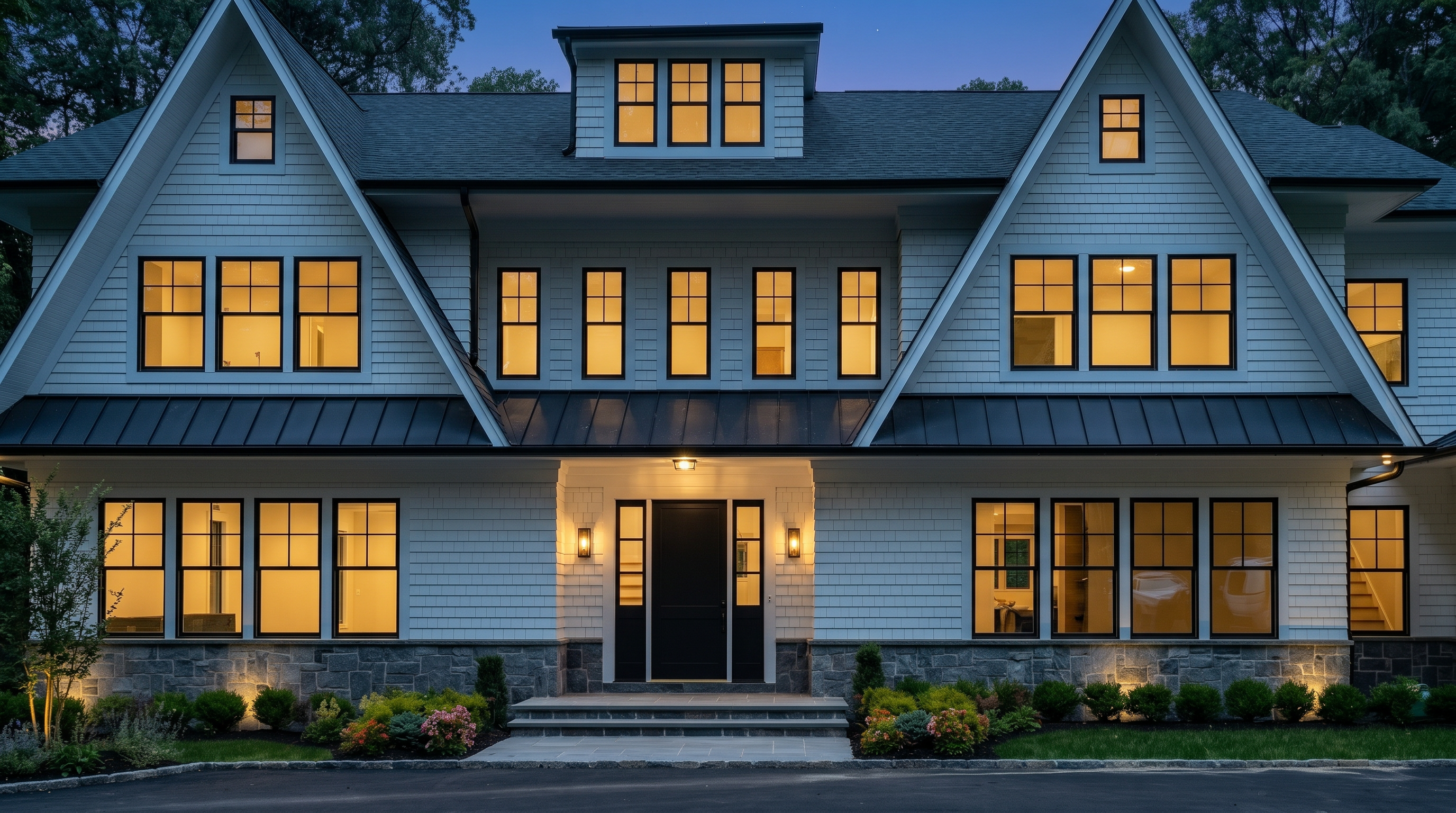 This is a striking front exterior view of a modern farmhouse-style residence at twilight, featuring symmetrical gables and abundant, warm-lit windows. The home showcases a blend of white shingle-style siding and classic stone masonry, complemented by a sleek dark metal roof and front entryway. Its sophisticated yet inviting design is highlighted by professional landscaping and exterior accent lighting, creating an elegant and high-end curb appeal.