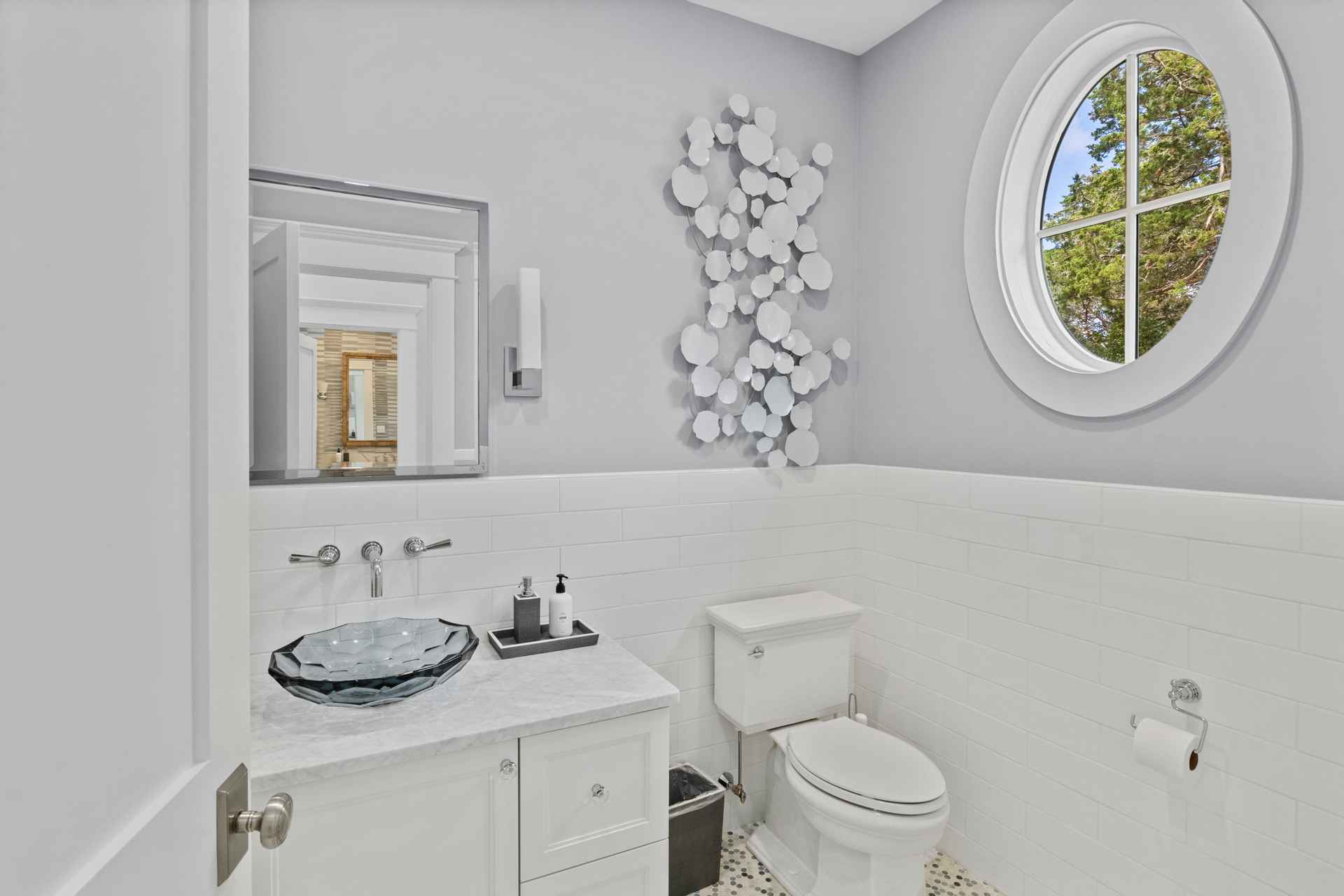 This is a well-lit guest bathroom featuring a unique glass vessel sink on a white vanity with marble countertop. The walls are partially tiled with white subway tiles, complemented by a light gray painted upper section. A round window provides natural light, and a modern, abstract wall sculpture adds a touch of elegance.