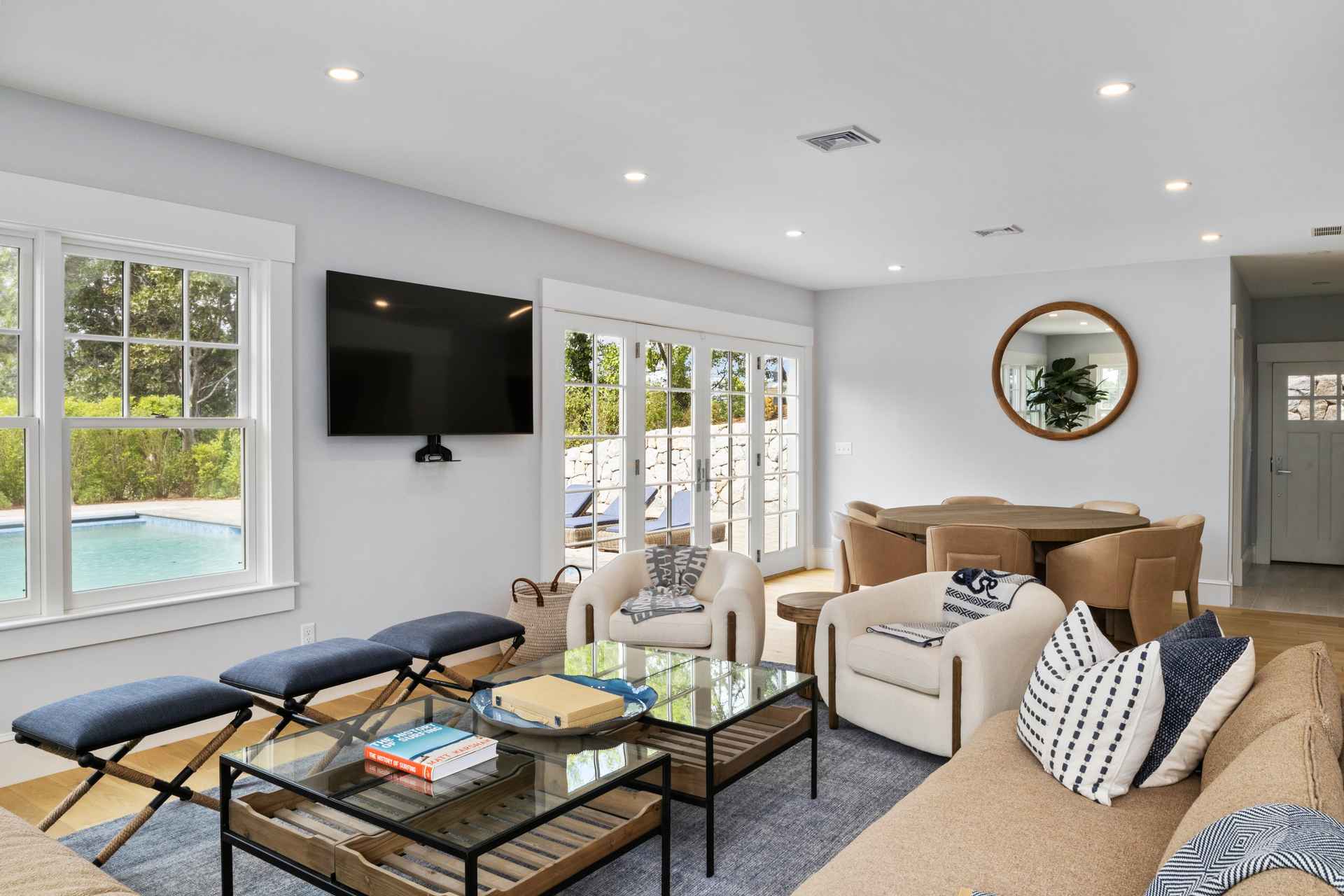 This is a bright and airy living room featuring a neutral color palette with pops of blue. The room is furnished with comfortable seating, including armchairs and a sectional sofa, arranged around a glass-topped coffee table. Large windows and glass doors provide ample natural light and views of the outdoor pool and patio area, enhancing the indoor-outdoor flow.