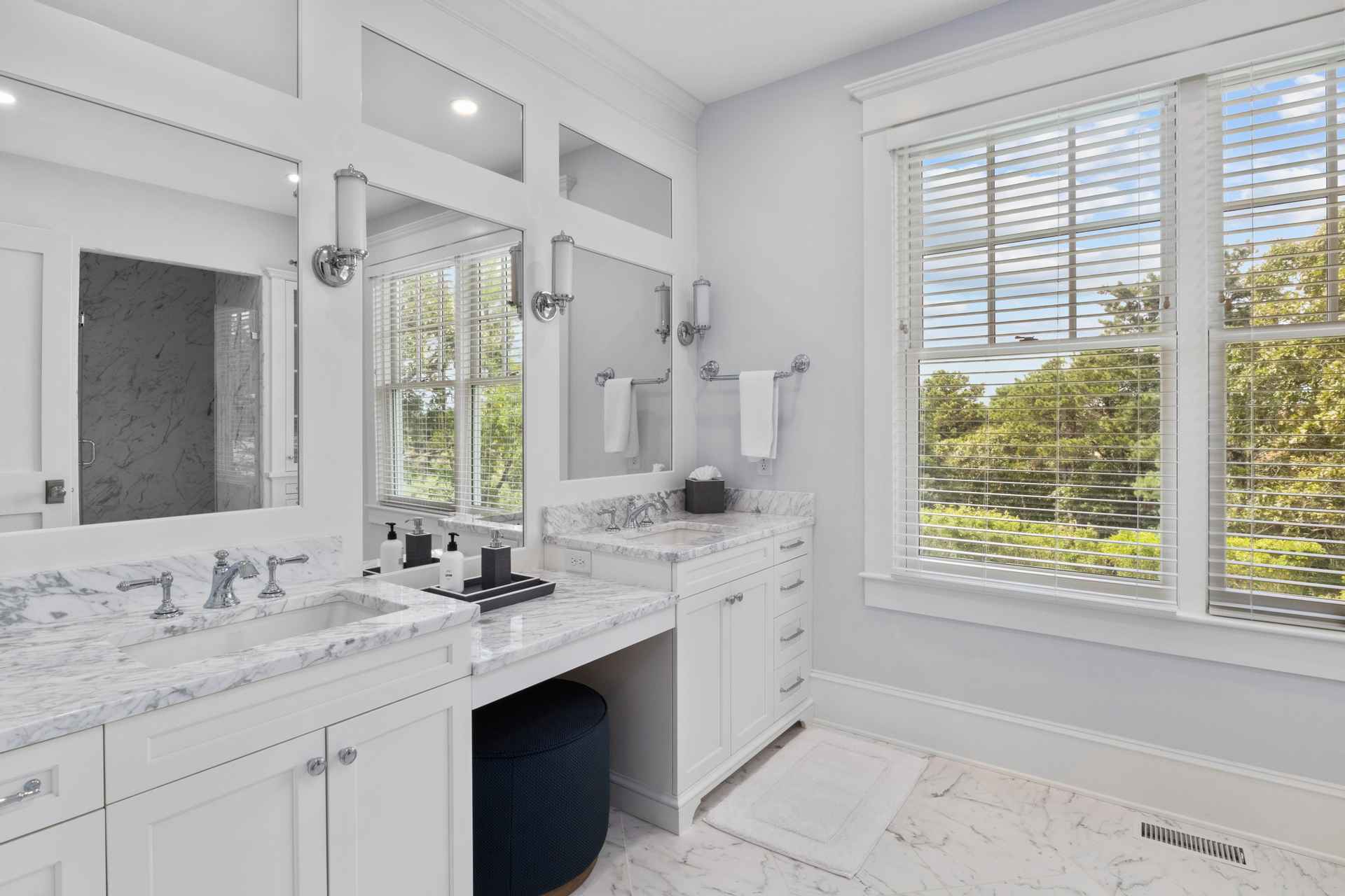 This is a bright and elegant primary bathroom featuring double vanities with white marble countertops and cabinetry. The room is well-lit with natural light from a large window with blinds, complemented by stylish sconces. The marble flooring and a navy blue ottoman add a touch of luxury to the space.