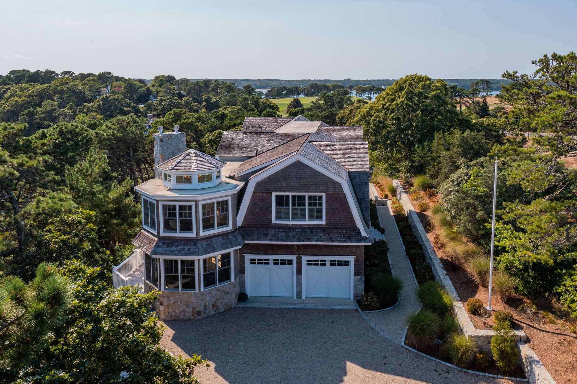 This aerial shot showcases a luxurious home nestled among lush greenery, offering a sense of privacy and tranquility. The house features a unique architectural design with a turret-like structure, a gambrel roof, and a two-car garage, all contributing to its distinctive curb appeal. A winding stone pathway and well-maintained landscaping add to the property's charm and upscale feel.