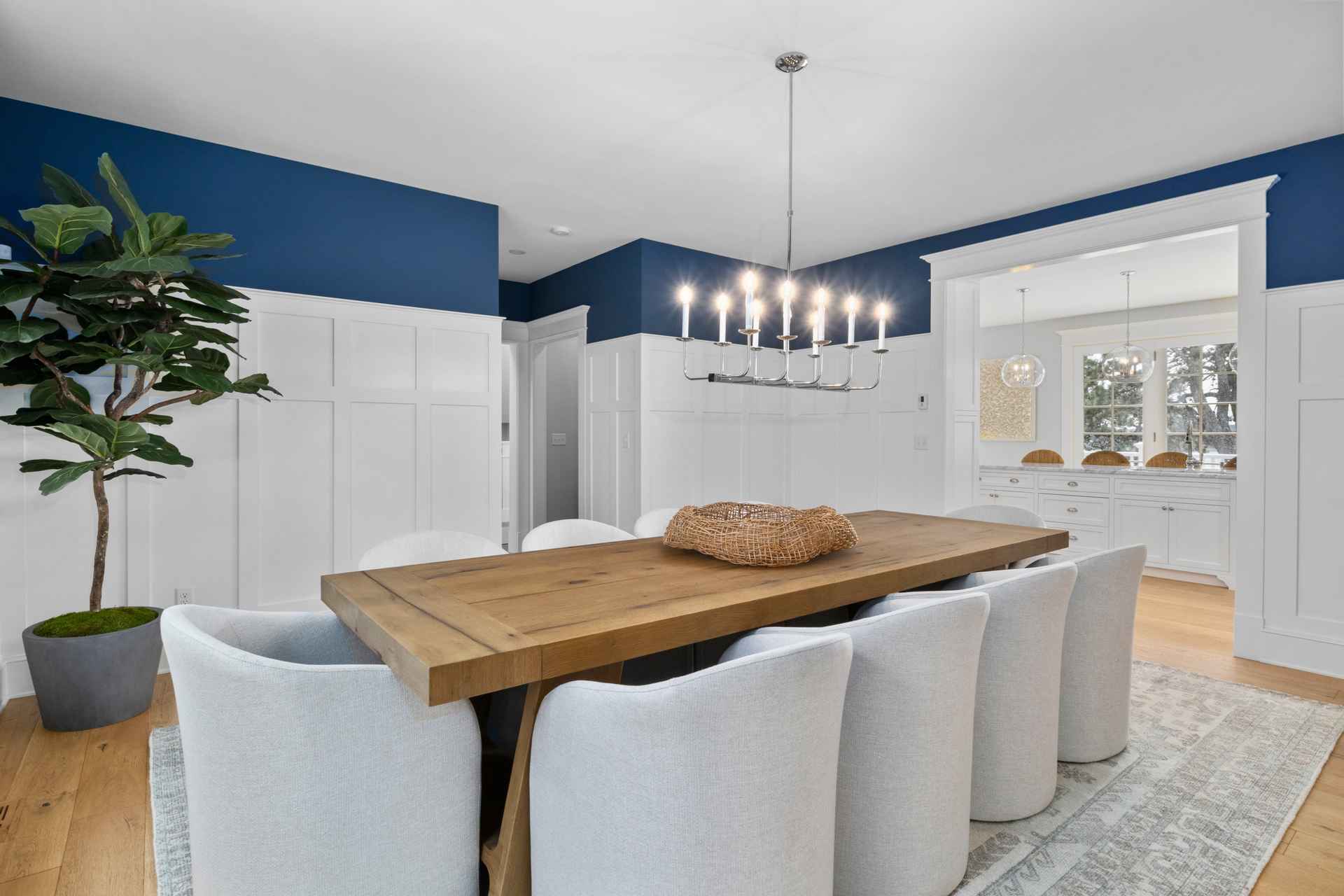 This is an interior shot of a dining room featuring a large wooden table surrounded by upholstered chairs. A modern chandelier hangs above the table, and the room is decorated with white wainscoting and a blue accent wall. The space feels bright and inviting, perfect for family meals and entertaining.