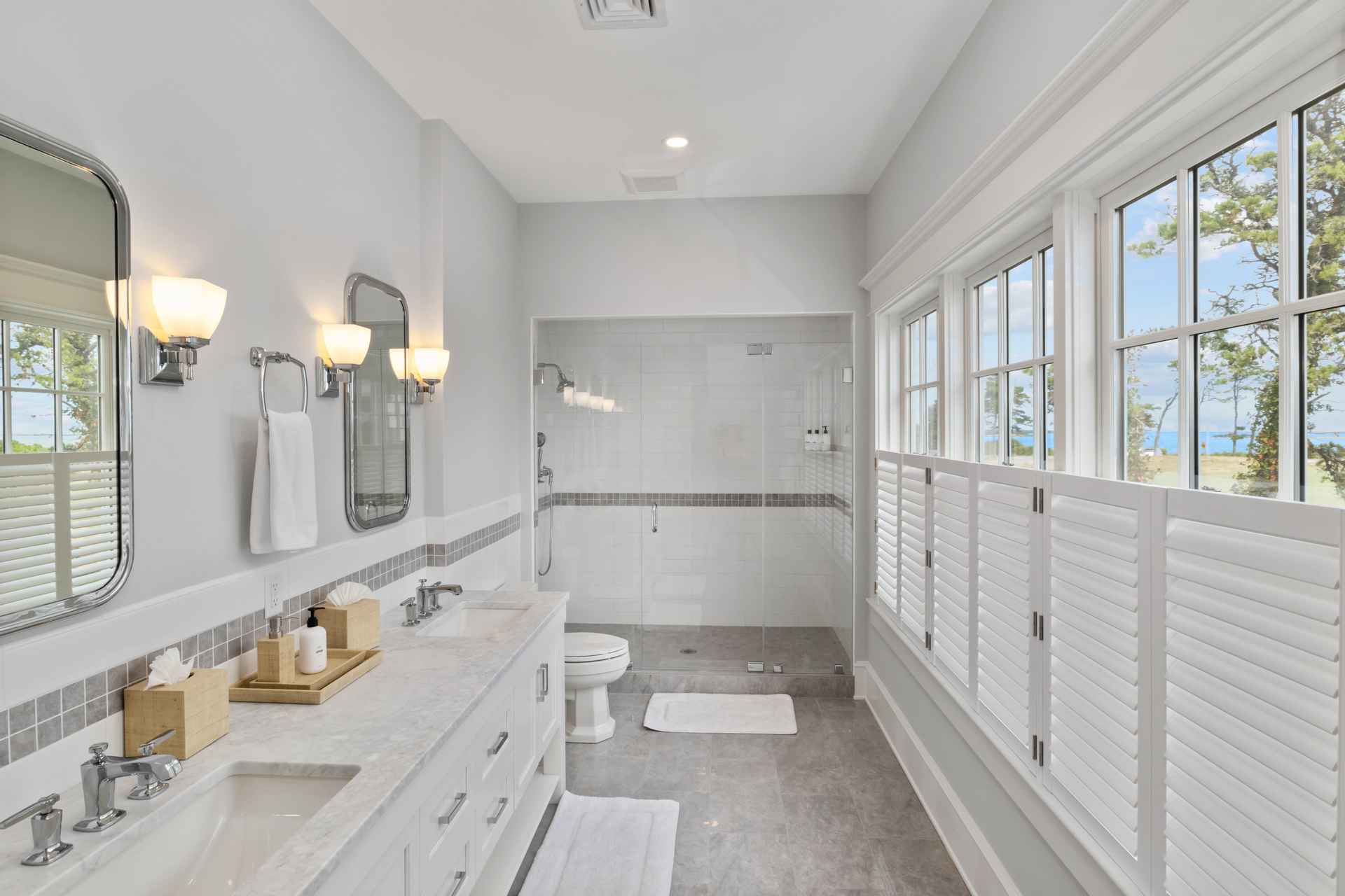 This is a bright and spacious primary bathroom featuring a double vanity with marble countertops and white cabinetry. The bathroom includes a glass-enclosed shower and large windows with white shutters, providing natural light and a view of the outdoors. The overall design is clean, modern, and luxurious, creating a spa-like atmosphere.
