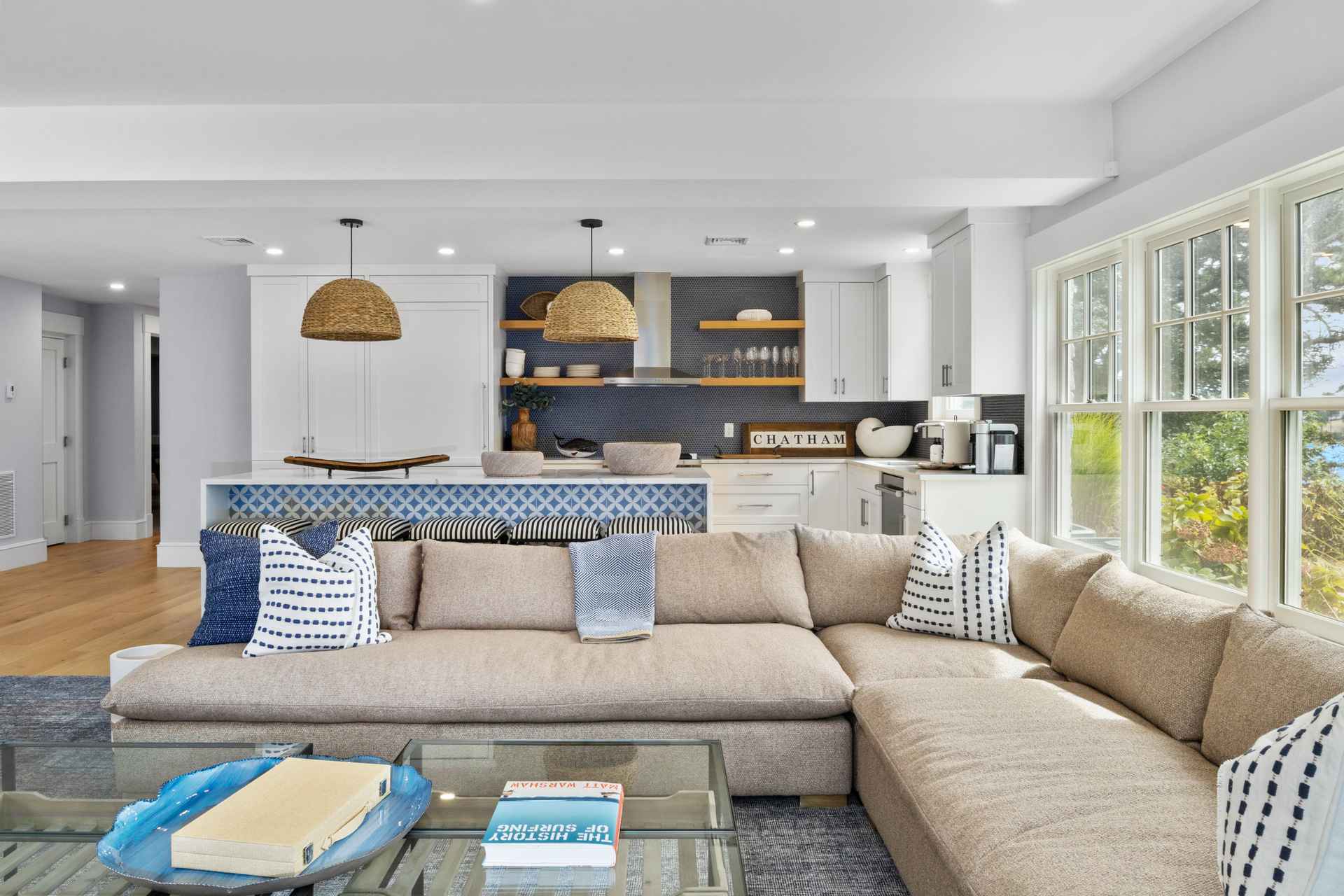 This is an interior shot of a well-lit living room that seamlessly blends into a modern kitchen. The living room features a large, comfortable sectional sofa adorned with decorative pillows, a glass coffee table with books and decorative items, and a neutral-toned rug. The kitchen area boasts white cabinetry, a blue tiled backsplash, and pendant lighting, creating a cohesive and stylish living space.
