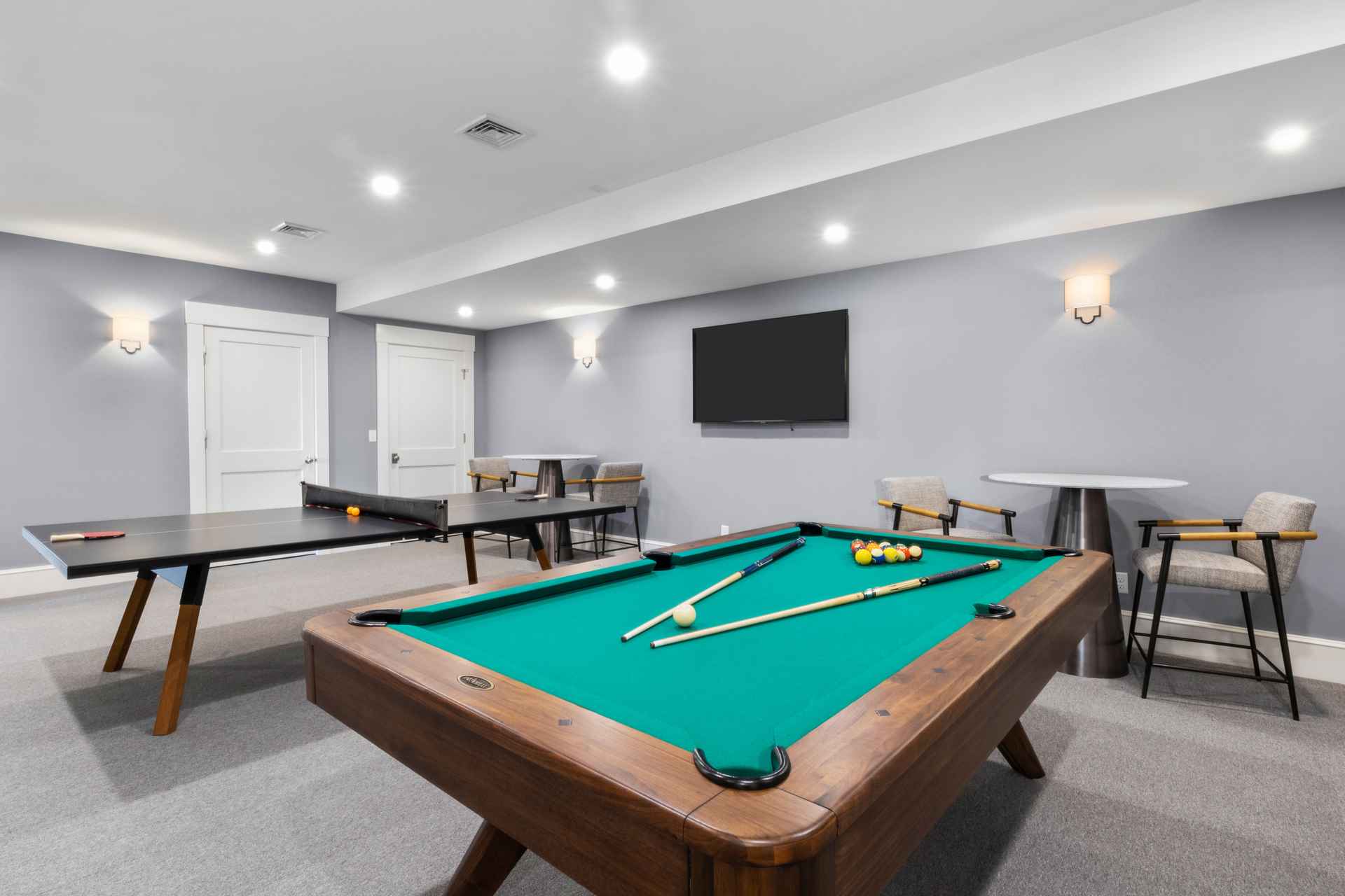 This interior shot showcases a recreational room featuring a pool table with cues and balls, a ping pong table, and a wall-mounted television. The room is painted in a neutral gray tone, complemented by recessed lighting and sconces, creating a relaxed and inviting atmosphere. Several chairs and small tables are arranged around the room, suggesting a space for leisure and entertainment.