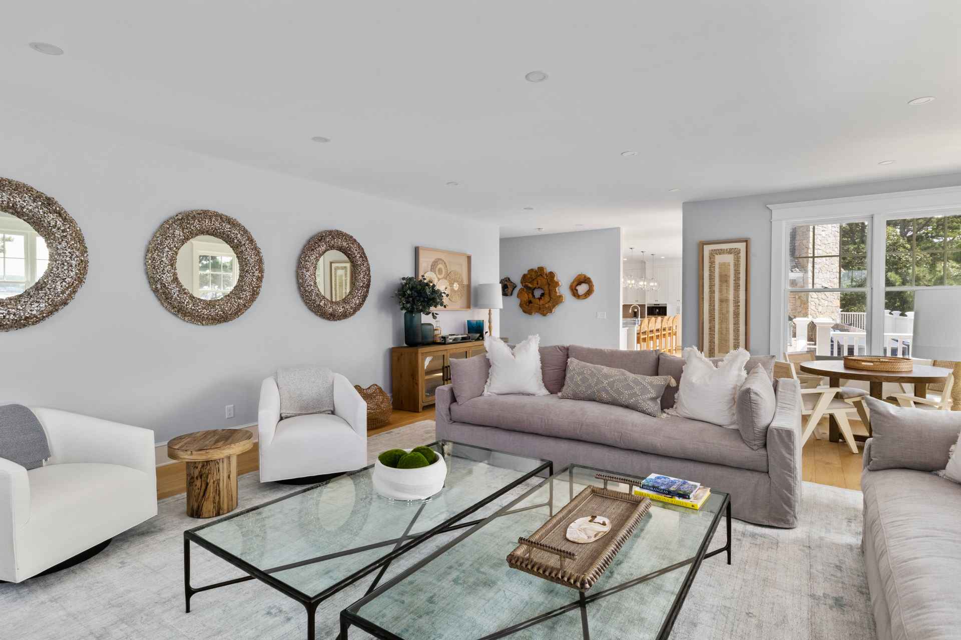 This is a bright and airy living room featuring a neutral color palette with pops of natural wood and greenery. The room is furnished with comfortable sofas, glass-topped coffee tables, and decorative mirrors on the wall. Large windows provide ample natural light, creating a welcoming and relaxing atmosphere.