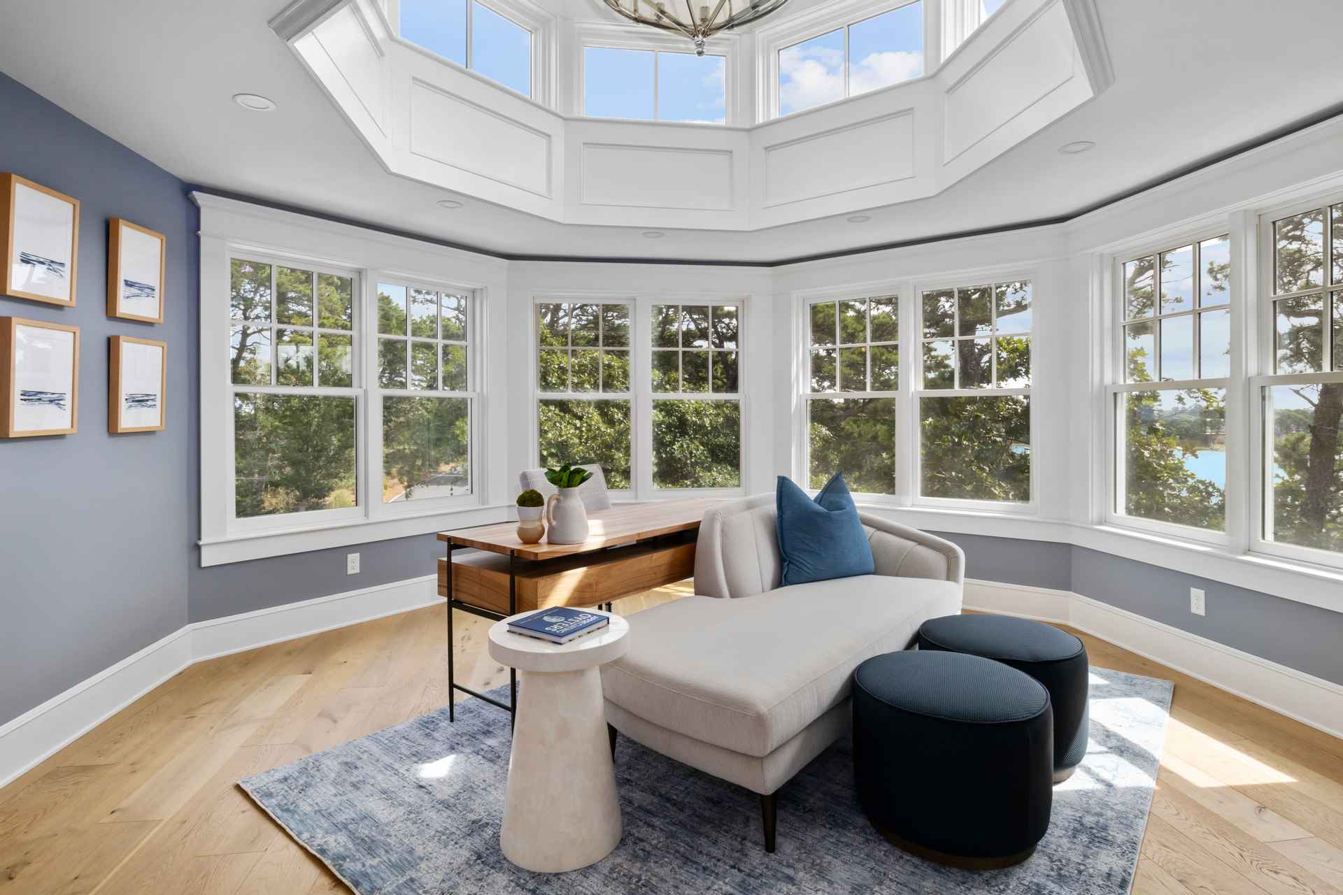 This is an interior shot of a bright and airy office or study. The room features a desk, a chaise lounge with blue pillows, and a round side table, all arranged on a blue patterned rug. Large windows surround the room, providing ample natural light and views of the surrounding greenery, creating a serene and productive workspace.