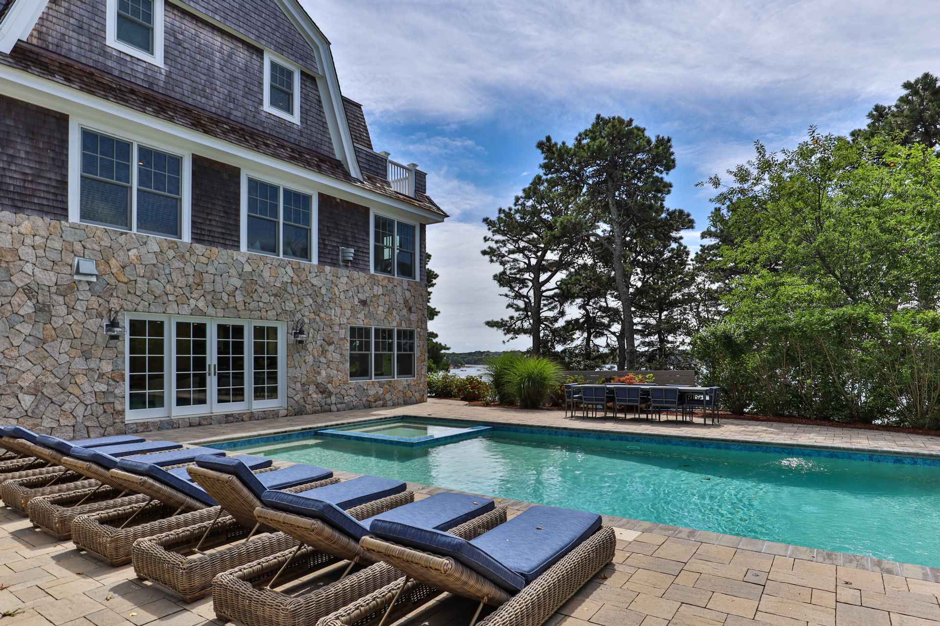This image showcases a luxurious backyard with a stunning pool and spa area. The pool is surrounded by a brick patio and features several lounge chairs with blue cushions. The house, constructed with stone and shingle siding, provides a beautiful backdrop, while mature trees and landscaping add privacy and elegance to the outdoor space.