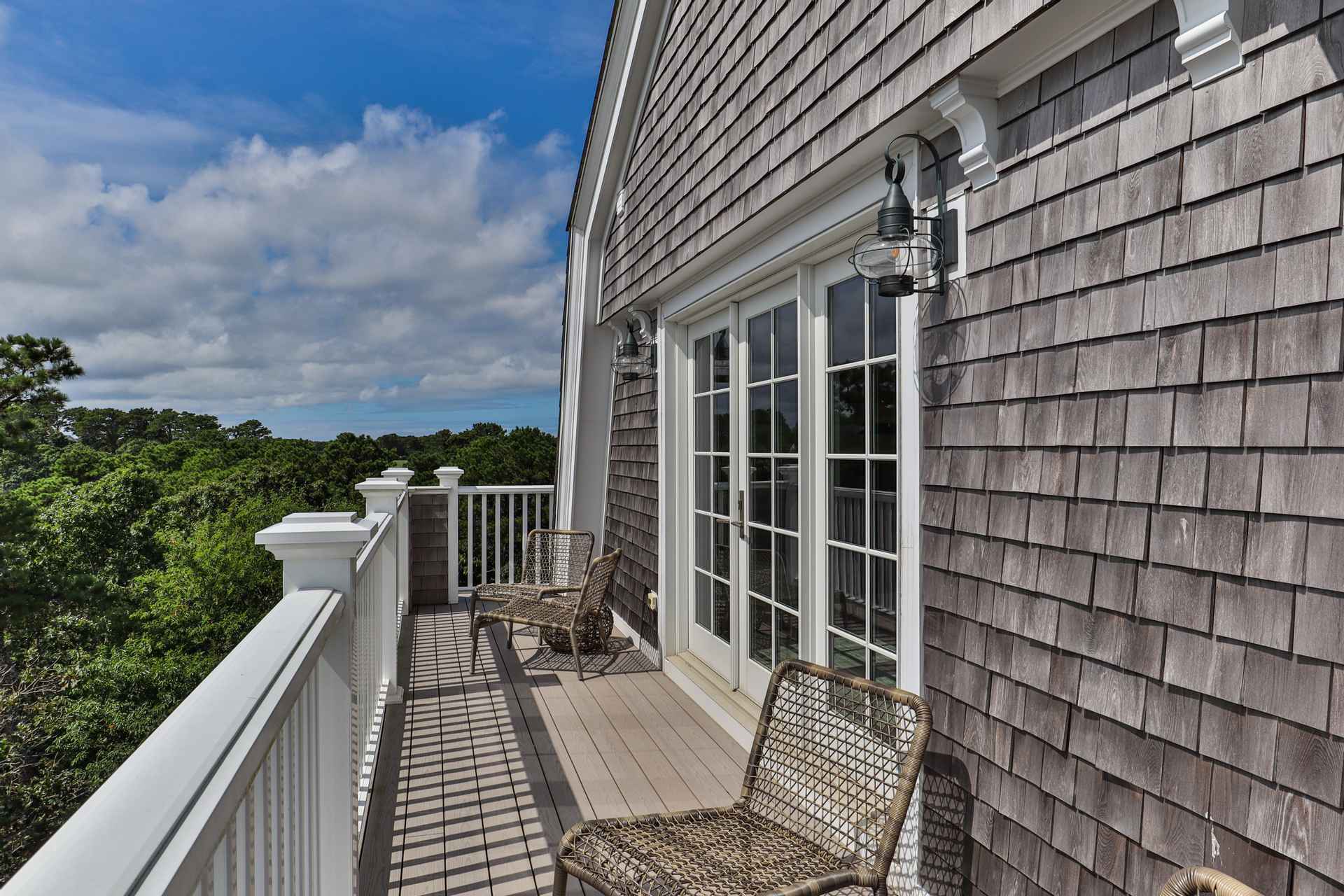 This image showcases a charming balcony with two woven chairs, offering a relaxing outdoor space. The balcony features a white railing and a wooden deck, complemented by the building's exterior with weathered gray shingles and classic lantern-style lighting fixtures. French doors provide access to the interior, creating a seamless transition between indoor and outdoor living.