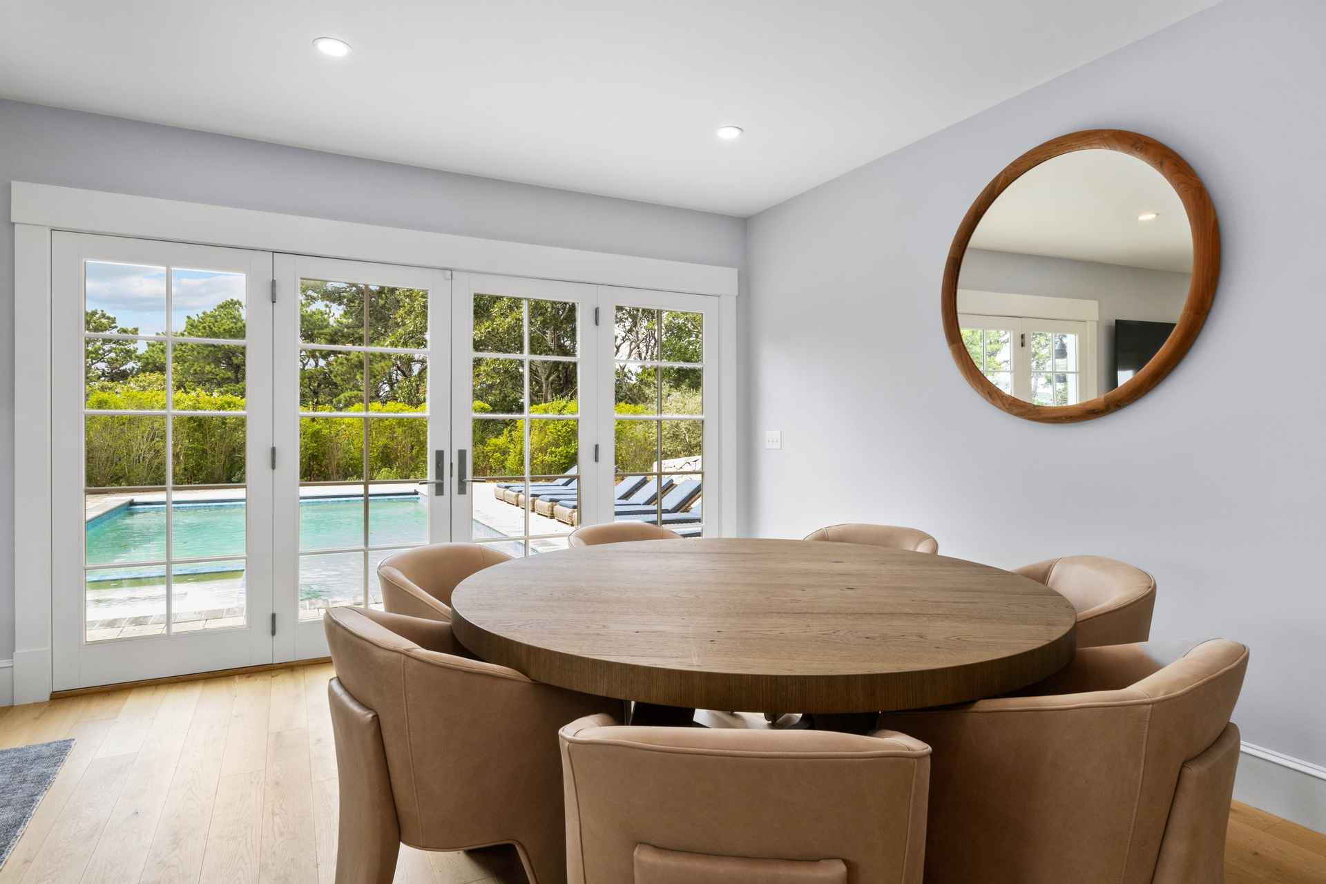 This is an interior shot of a dining room featuring a round wooden table surrounded by upholstered chairs. Large French doors offer a view of a pool and outdoor lounge area, bringing natural light into the space. A round mirror hangs on the wall, adding depth and style to the room.