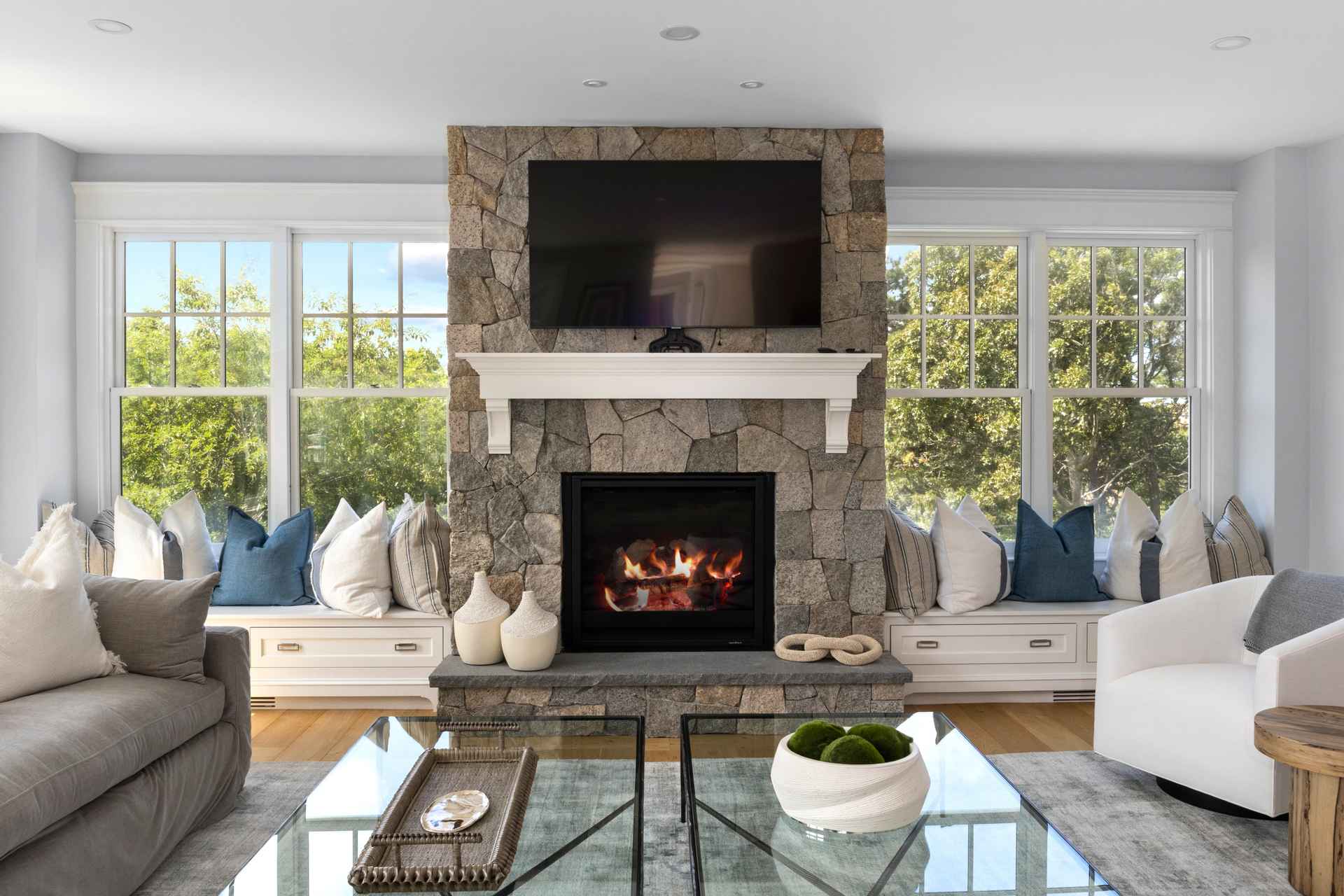This is an interior shot of a living room featuring a stone fireplace with a mounted television above a white mantel. The room is well-lit with natural light streaming through large windows on either side of the fireplace, which have built-in window seats with decorative pillows. The furniture includes a gray sofa, a white armchair, and glass-topped coffee tables, creating a comfortable and inviting atmosphere.