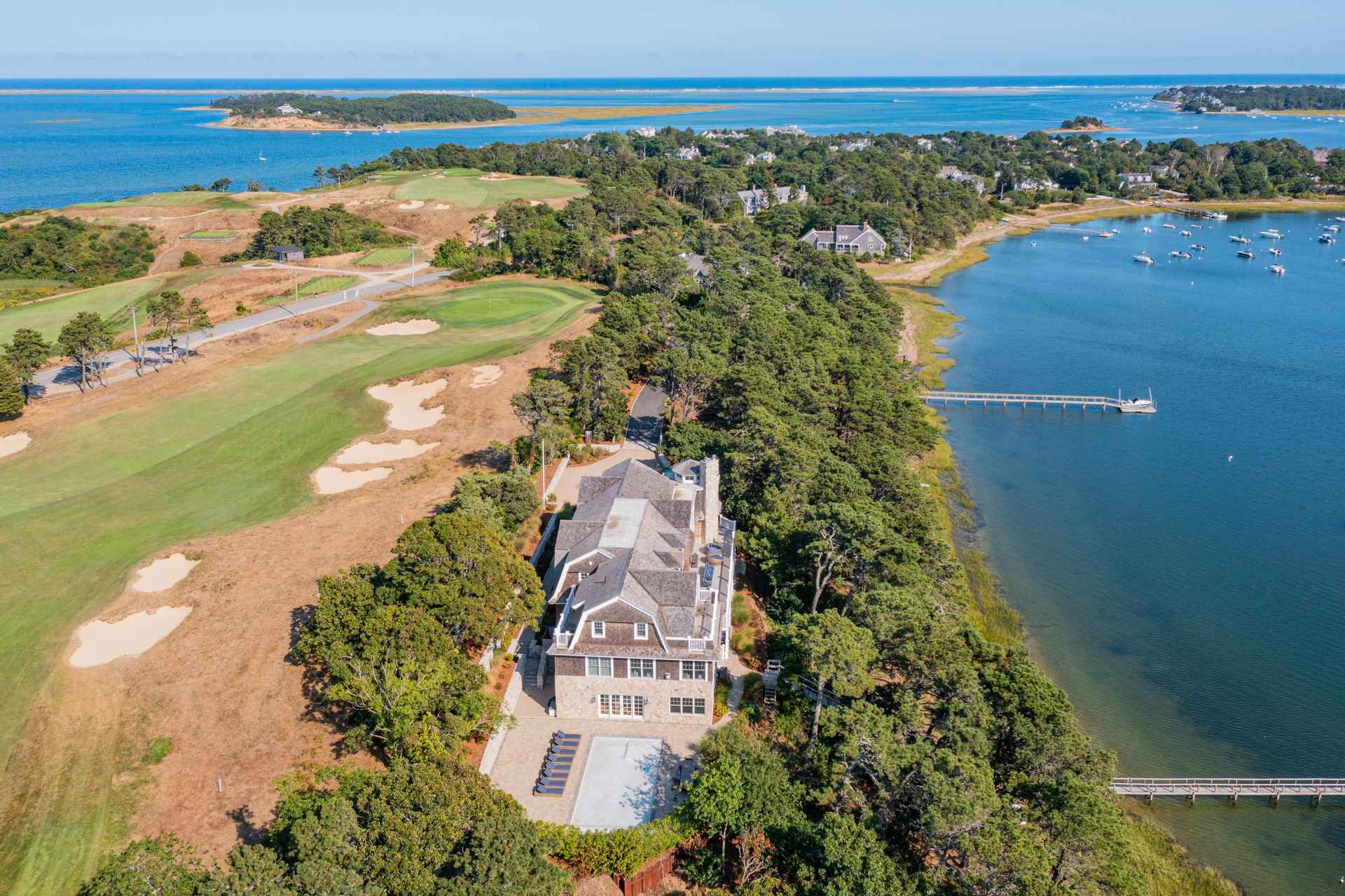 This aerial view showcases a luxurious property nestled between a golf course and a waterfront. The house features a stone facade, a shingle roof, and a pool area. The surrounding landscape includes lush trees and a private dock, creating an exclusive and serene setting.