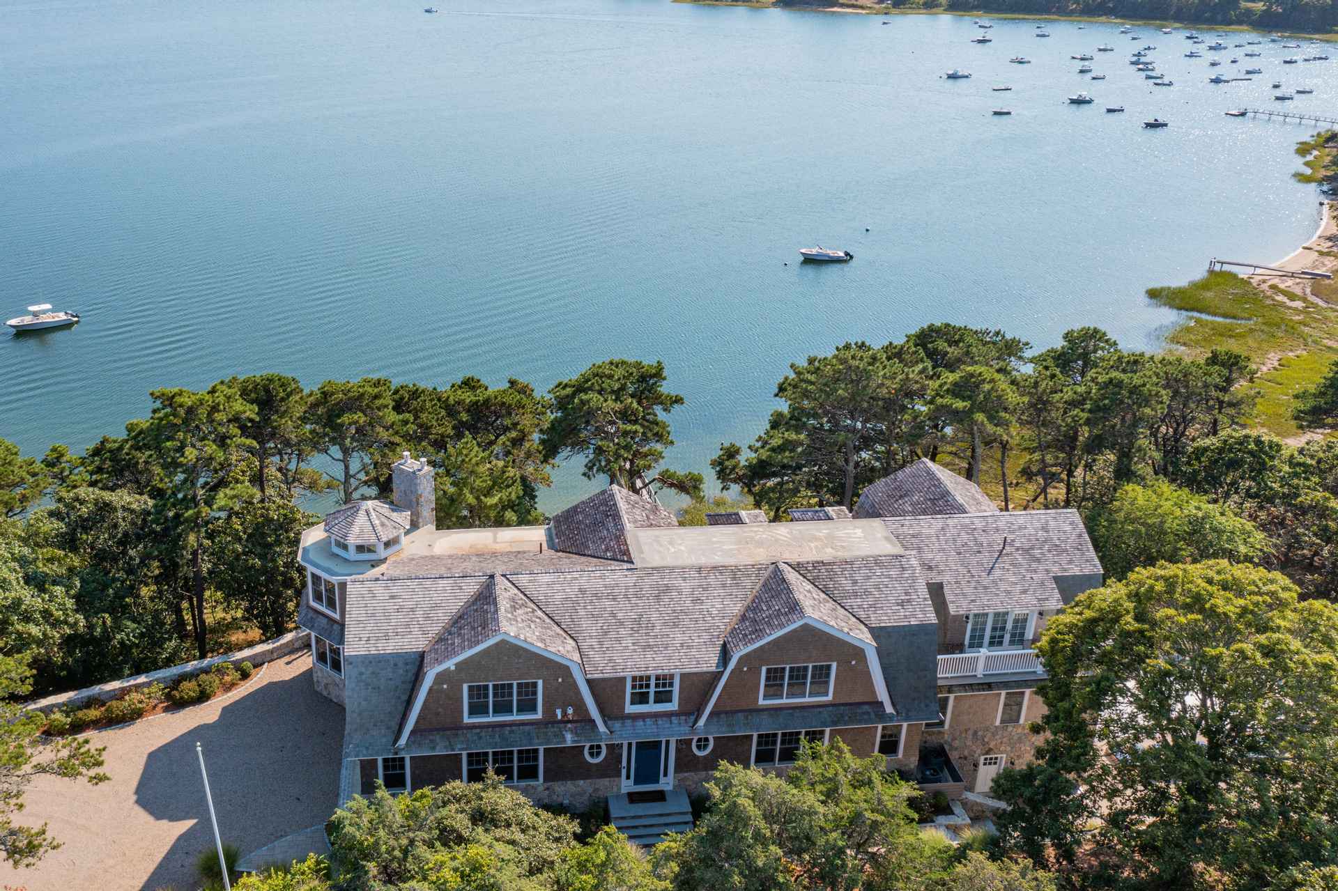 This aerial shot showcases a large, luxurious waterfront home with a complex roofline, stone and wood siding, and a turret-like structure. The property is surrounded by mature trees, offering privacy, and overlooks a serene bay dotted with boats. The overall impression is one of high-end coastal living.
