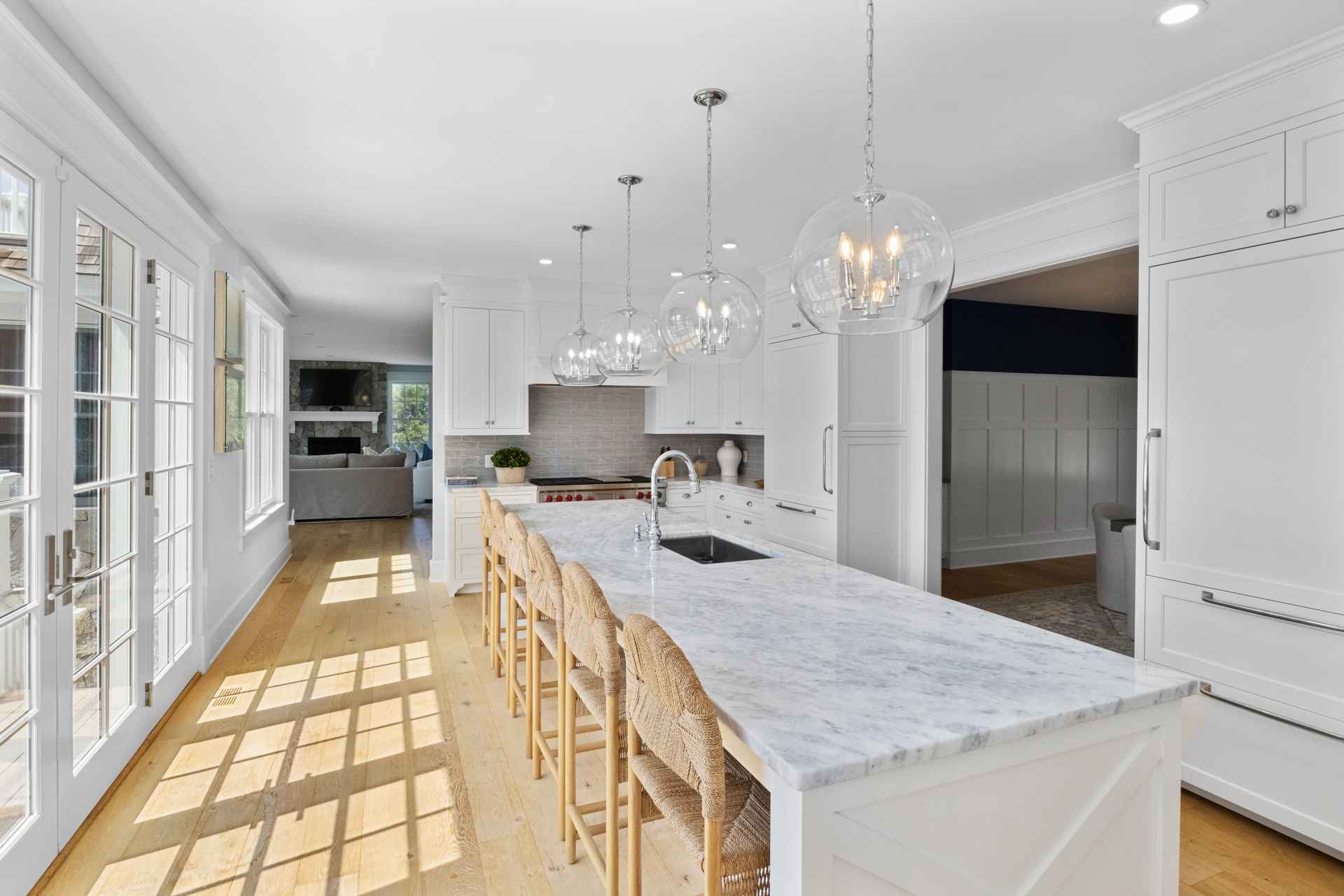 This is a bright and spacious kitchen featuring a large marble island with seating, white cabinetry, and stainless steel appliances. The kitchen is well-lit with natural light from the windows and pendant lighting above the island. The open floor plan leads to a living area, creating a welcoming and modern space.