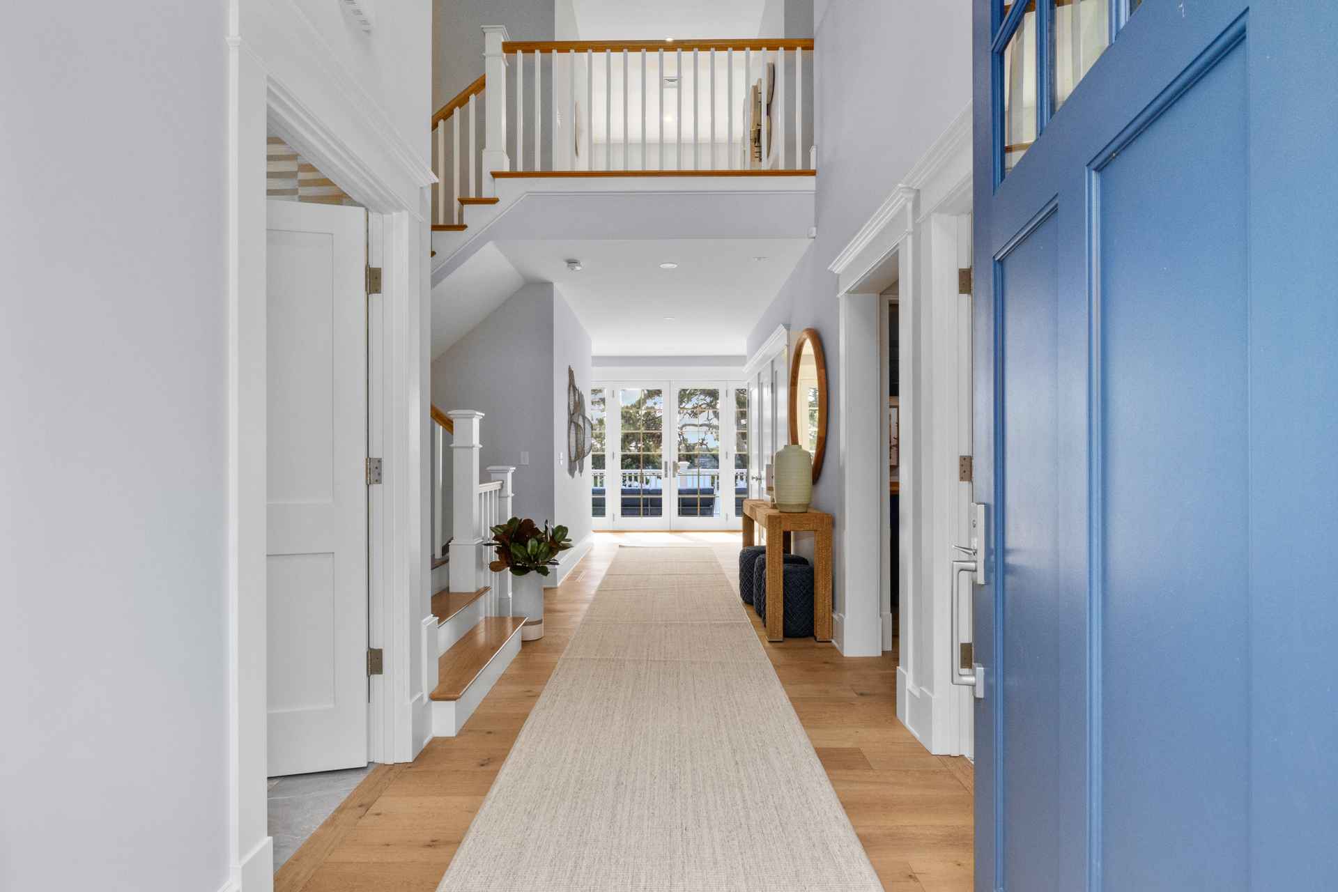 This is an interior shot of a bright and airy hallway featuring hardwood floors and a long runner rug. A blue front door is partially open on the right, and stairs lead to a second-floor landing with white railings. Natural light floods the space through a set of glass doors at the end of the hallway, creating an inviting and spacious feel.