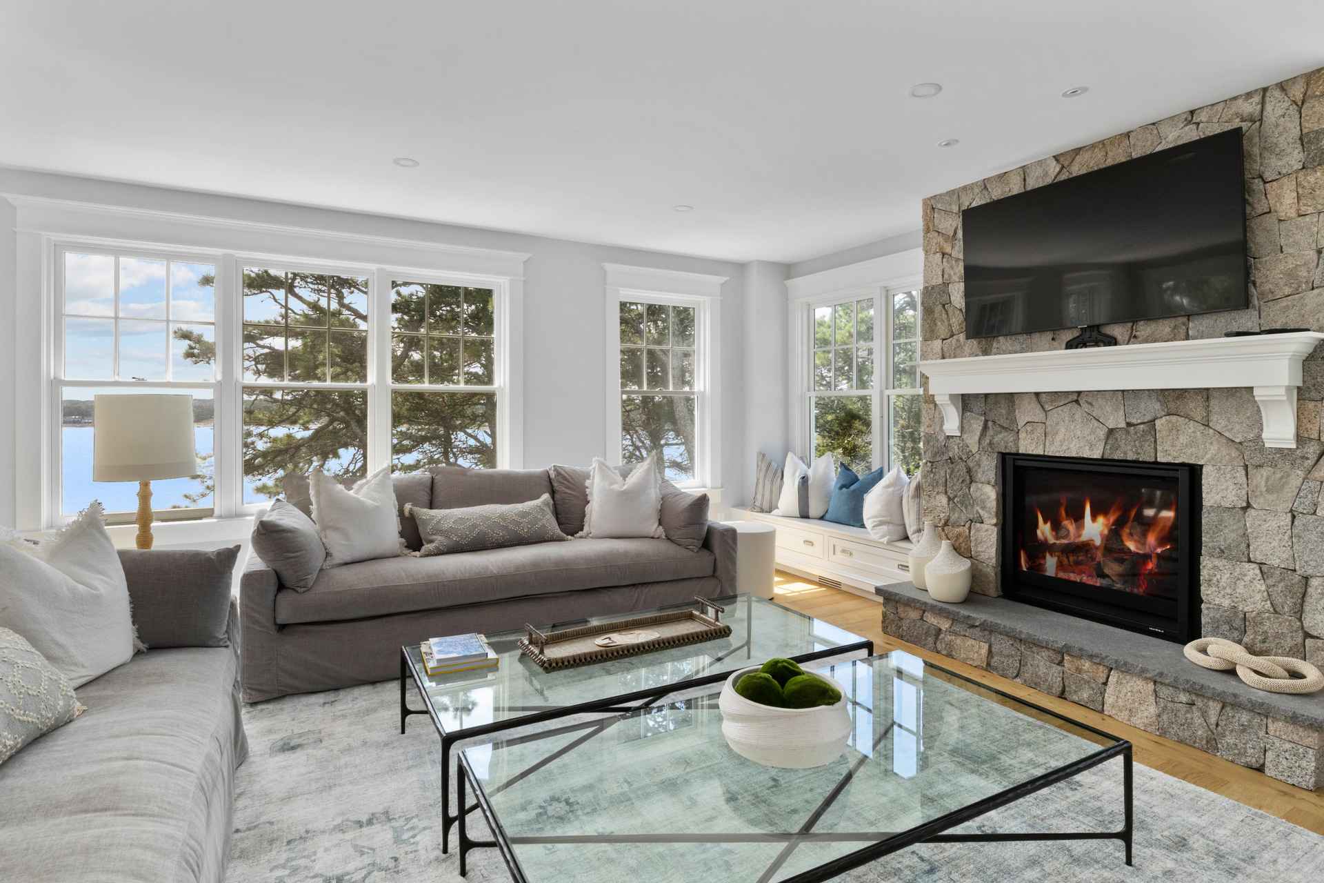 This is a bright and airy living room featuring a stone fireplace with a mounted television above. The room is furnished with comfortable gray sofas and glass coffee tables, complemented by large windows offering scenic views. The overall design creates a cozy yet elegant atmosphere, perfect for relaxation and entertaining.