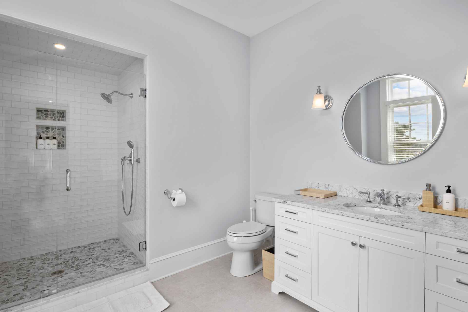 This is a well-lit primary bathroom featuring a glass-enclosed shower with white subway tile and pebble flooring. A white vanity with a marble countertop and a round mirror are prominent, complemented by chrome fixtures and sconce lighting. The overall aesthetic is clean, modern, and luxurious.