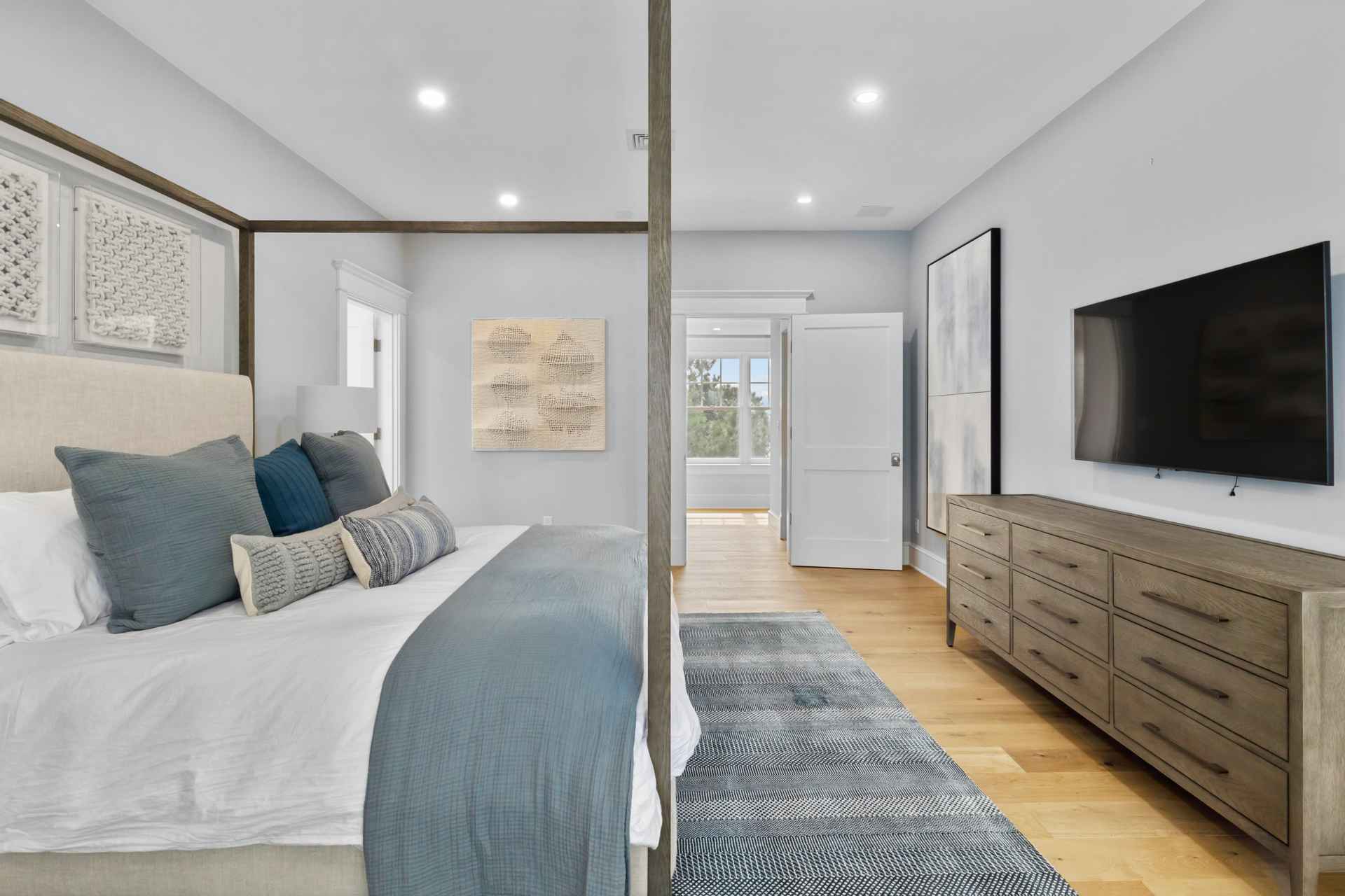This is a bright and airy primary bedroom featuring a four-poster bed with blue and gray pillows and a blue throw. The room has light hardwood floors, a gray dresser with a large television mounted above it, and modern artwork on the walls. A doorway leads to another room, suggesting an en-suite bathroom or dressing area.