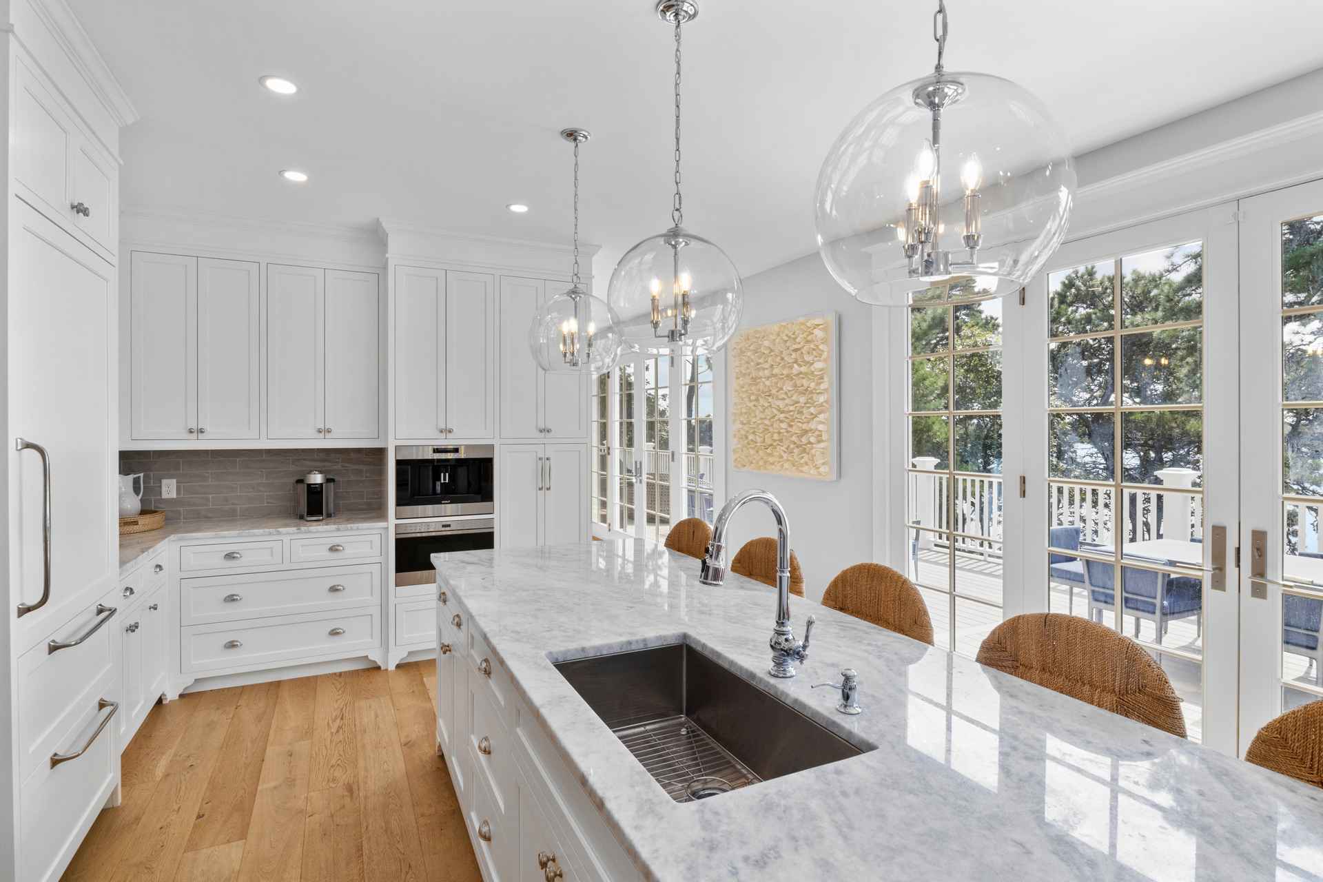 This is a bright and airy kitchen featuring white cabinetry, marble countertops, and stainless steel appliances. A large island with a sink and seating area is the focal point, illuminated by three glass pendant lights. French doors lead to an outdoor deck, offering a view of the surrounding landscape.