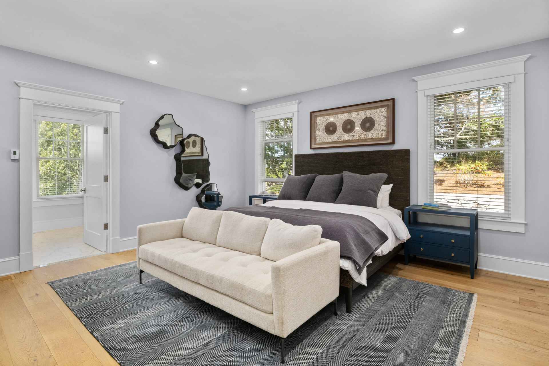 This is a well-lit primary bedroom featuring a neutral color palette with light blue walls and hardwood floors. A large bed with a dark headboard is the focal point, complemented by matching nightstands and artwork. A modern sofa sits at the foot of the bed on top of a patterned rug, adding a touch of elegance and comfort to the space.