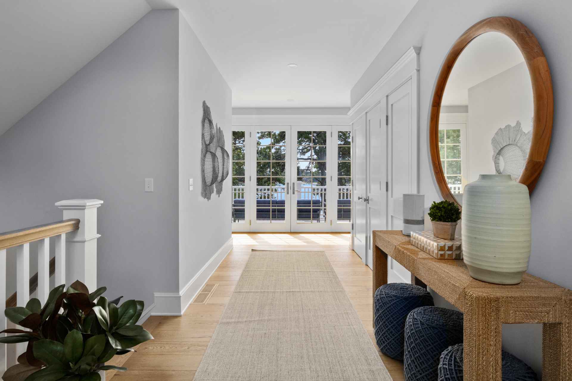 This is an interior shot of a bright and airy hallway. The hallway features light hardwood floors, light gray walls, and white trim. A long runner rug leads towards a set of French doors that open to an outdoor area with a view of the water. A console table with decorative items and a round mirror adds a touch of elegance to the space.