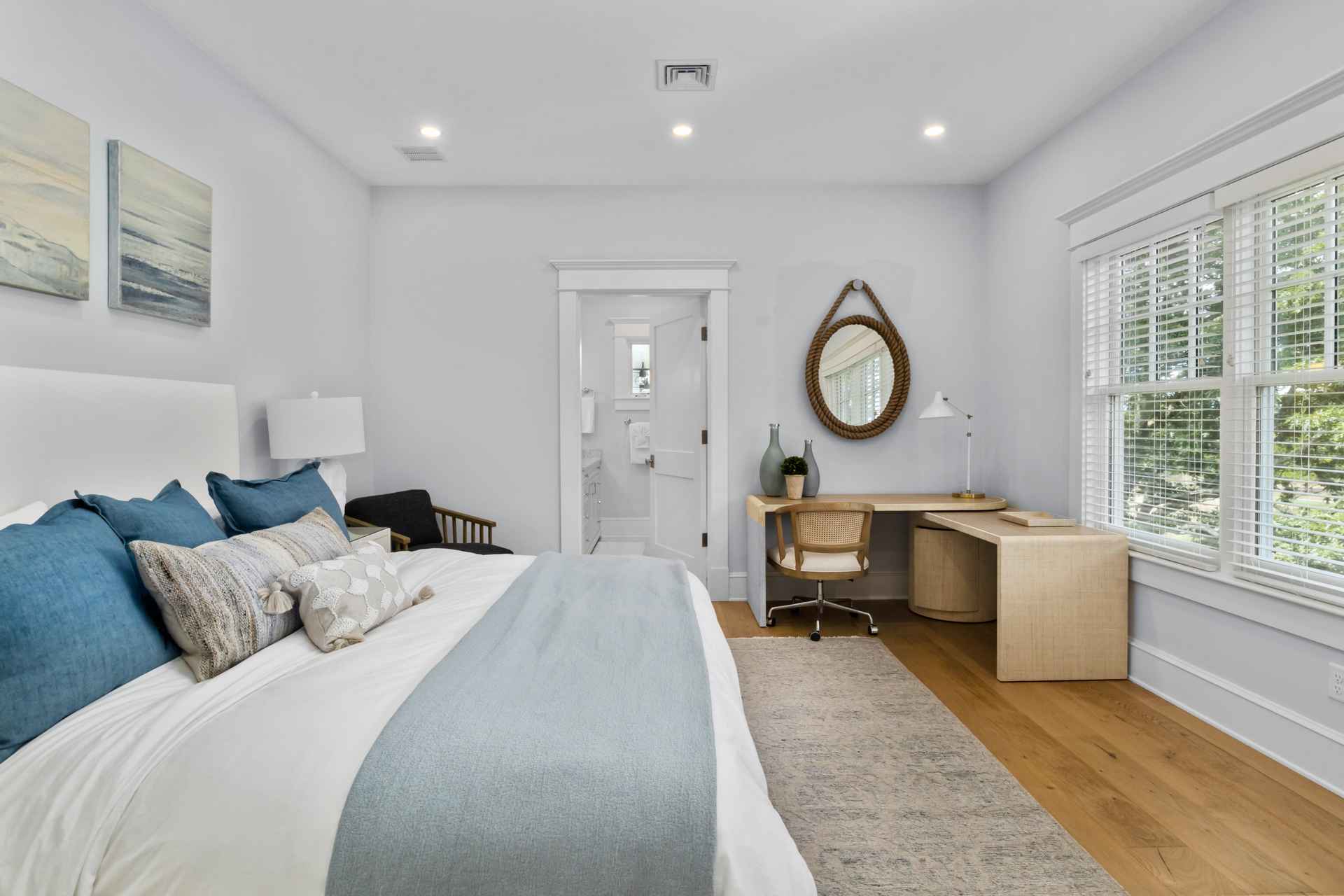 This is a bright and airy primary bedroom featuring a large bed with blue and patterned pillows, a light blue throw, and white linens. A desk with a woven chair sits near a window with blinds, and a round mirror hangs above the desk. The room has light gray walls, hardwood floors, and recessed lighting, creating a serene and inviting atmosphere.