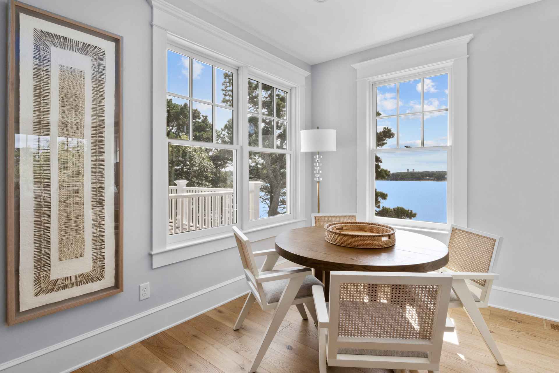 This is a bright and airy dining room featuring a round wooden table surrounded by four white chairs with woven backs. Natural light floods the space through two large windows, offering a view of the water and trees. A large framed artwork hangs on the wall, adding a touch of sophistication to the room's coastal aesthetic.