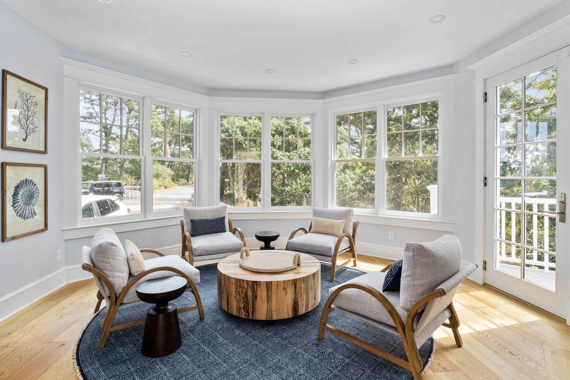 This is an interior shot of a sunroom or living room area featuring a circular seating arrangement. Four armchairs with light-colored cushions are positioned around a round, wooden coffee table on a blue patterned rug. Large windows surround the space, providing ample natural light and views of the outdoors, while artwork adorns the wall, creating a cozy and inviting atmosphere.