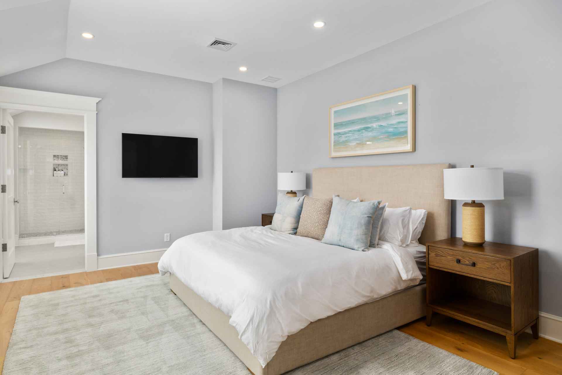 This is a well-lit primary bedroom featuring a king-size bed with a beige headboard and white linens, complemented by blue and patterned throw pillows. A wooden nightstand with a lamp sits beside the bed, and a framed ocean-themed artwork hangs above the headboard. A flat-screen TV is mounted on the wall opposite the bed, and a doorway leads to an en-suite bathroom, creating a serene and inviting atmosphere.
