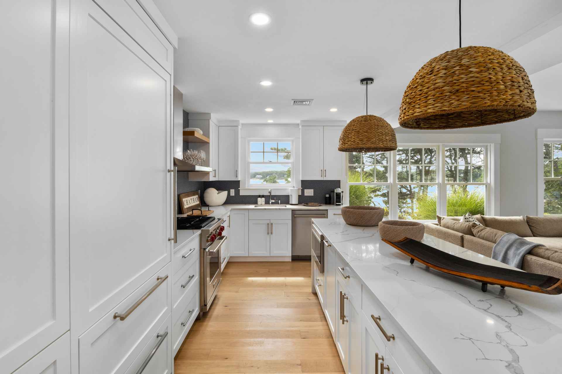 This is a bright and modern kitchen featuring white cabinetry, stainless steel appliances, and a large island with a marble countertop. Woven pendant lights hang above the island, adding a touch of natural texture. The kitchen offers a view of the outdoors through a window, creating a serene and inviting atmosphere.